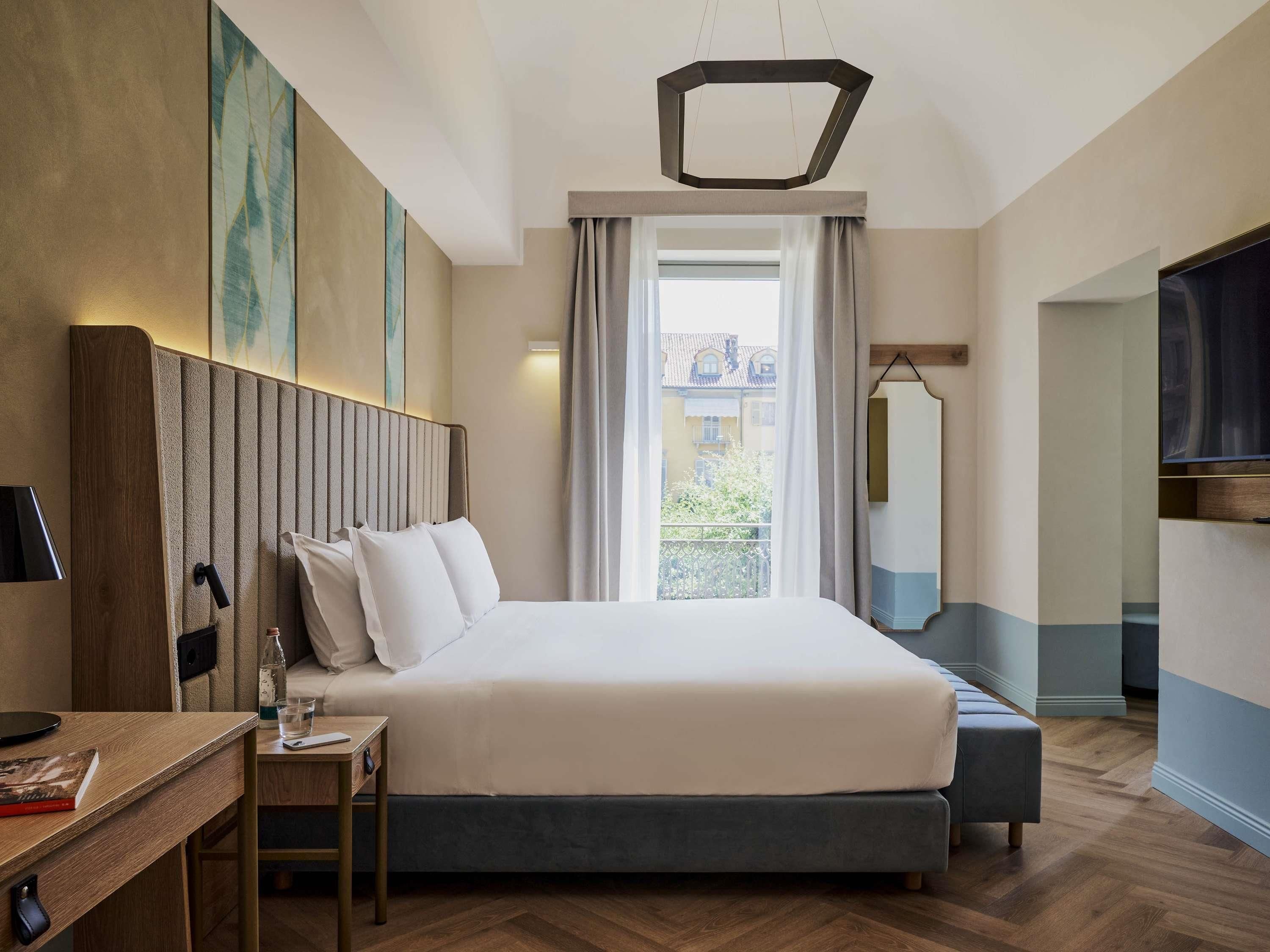 A hotel room with a large bed, a desk with a lamp, a TV, and a view of buildings through the window.
