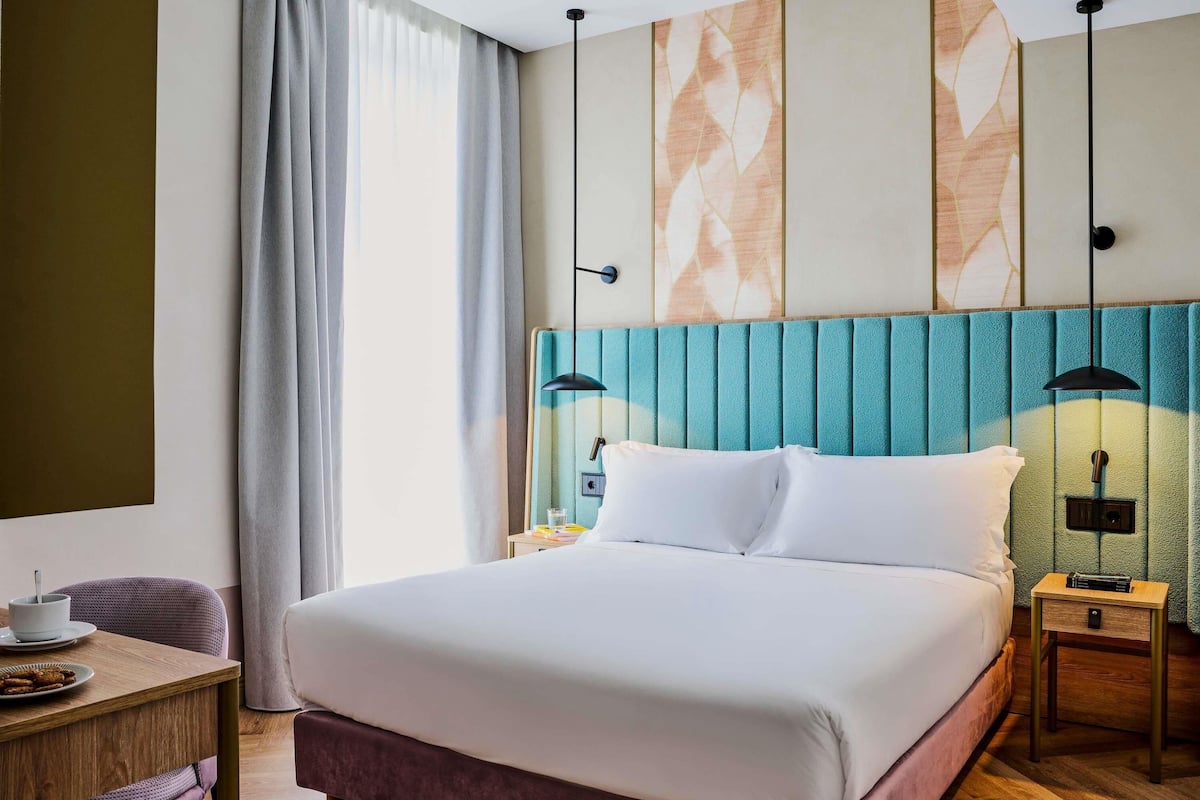 A hotel room with a large bed, a bedside table with a lamp, a chair, and a view of the outside through the window.