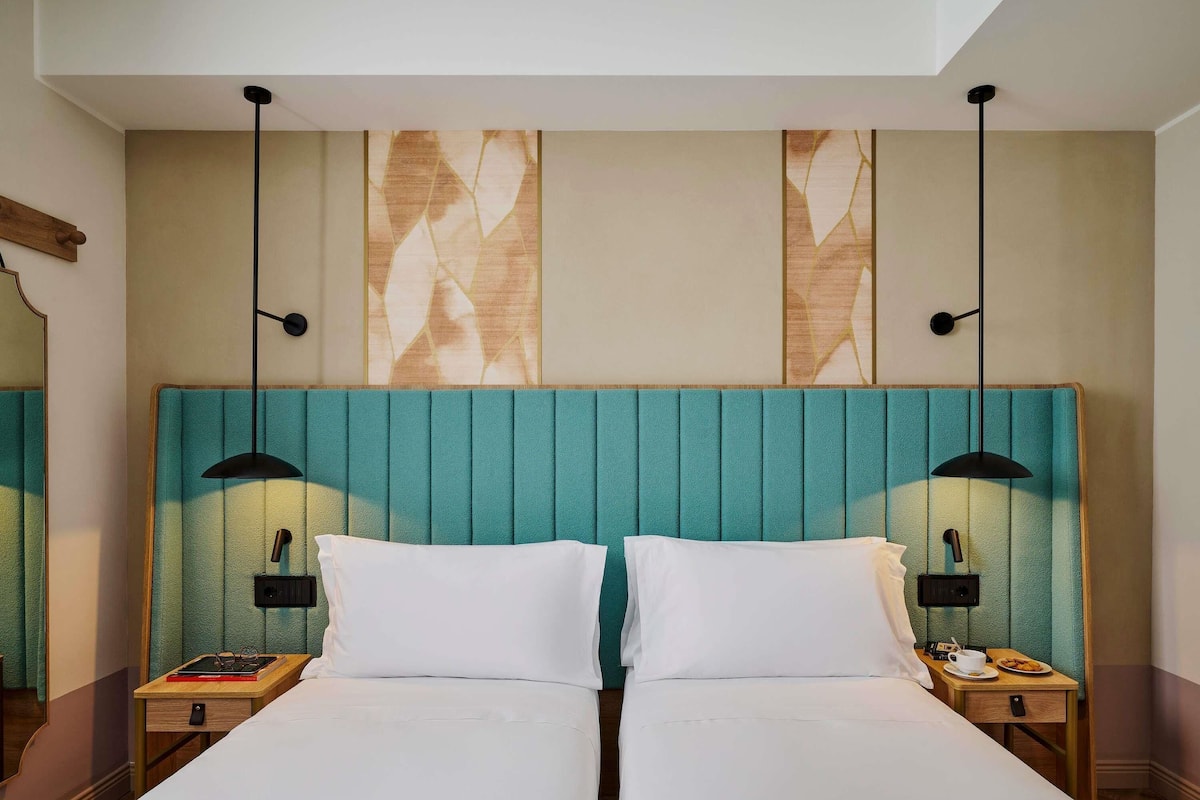 A double bed with a teal headboard, two bedside tables with lamps, and a mirror on the left wall.