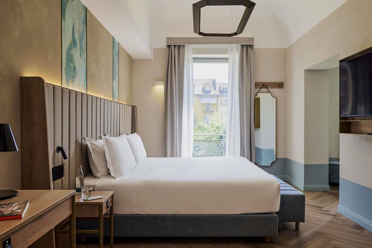A hotel room with a large bed, a desk with a lamp, a TV, and a view of buildings through the window.