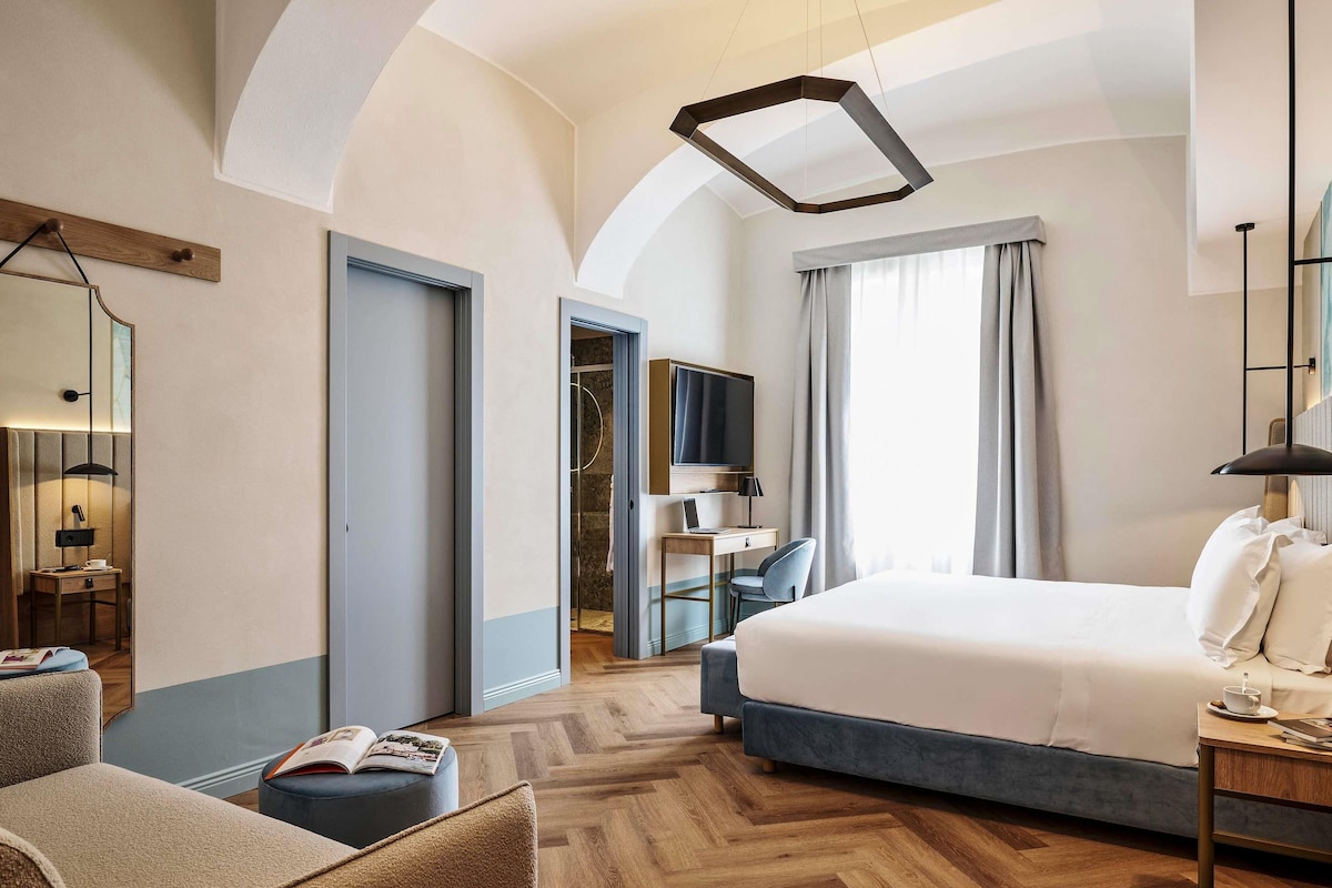 A modern hotel room with a large bed, a desk with a laptop, a chair, and a small seating area with a book.