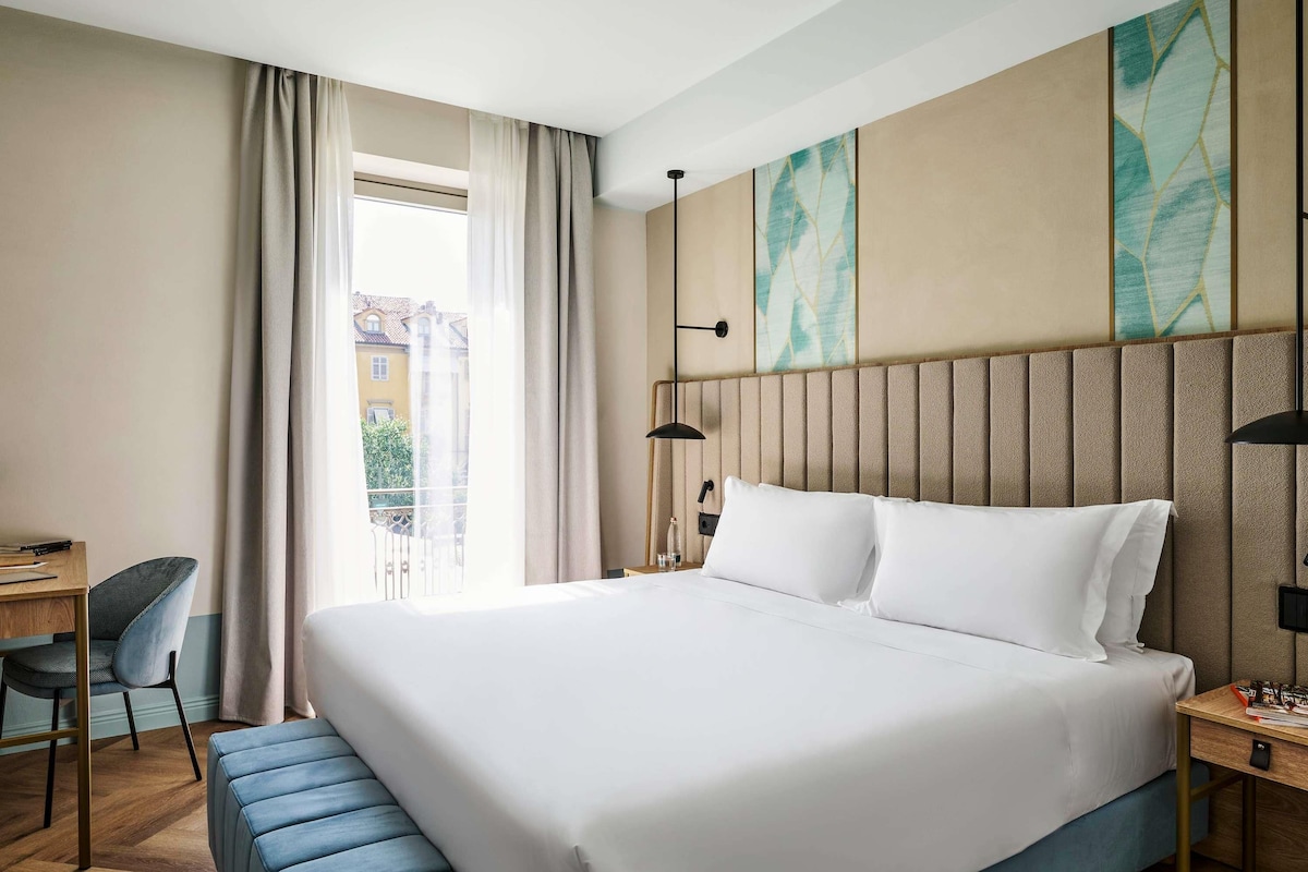 A hotel room with a large bed, a desk, a chair, and a view of the outside.