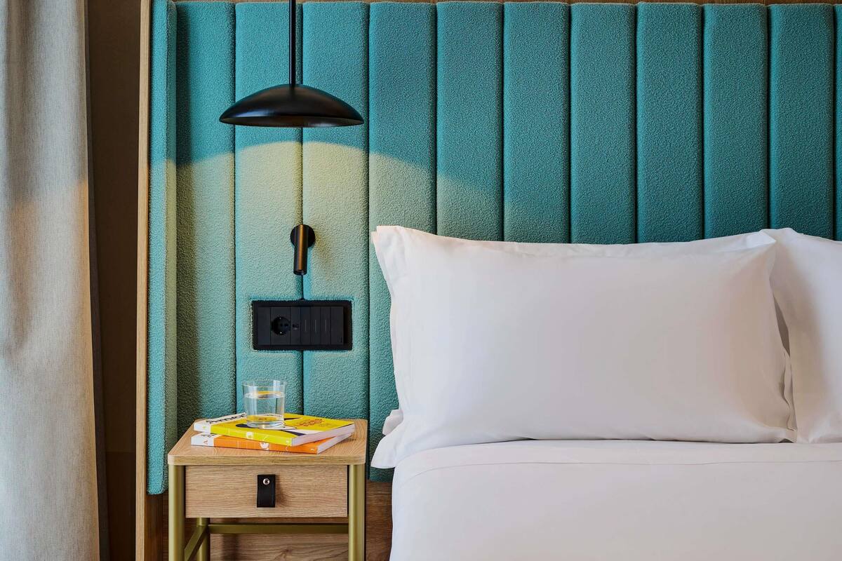 A neatly made bed with white linens, a bedside table with a lamp, and a wall panel with a switch.
