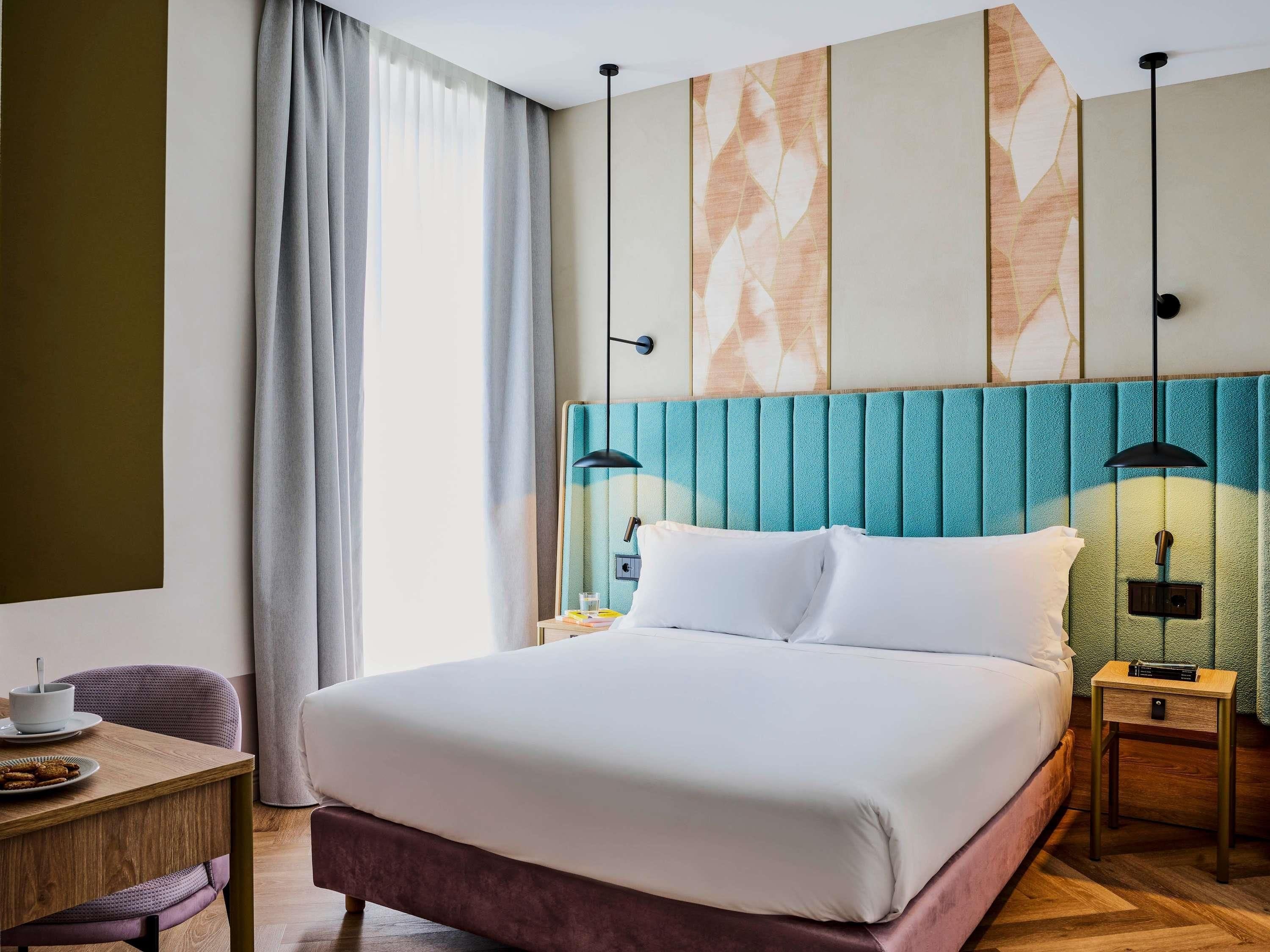 Ein Hotelzimmer mit einem großen Bett, einem Nachttisch mit Lampe, einem Sessel und Blick ins Freie durch das Fenster.