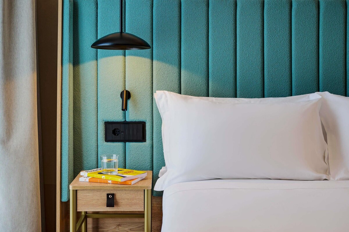 Un lit bien fait, avec du linge de lit blanc, une table de chevet avec une lampe et un panneau mural avec un interrupteur.