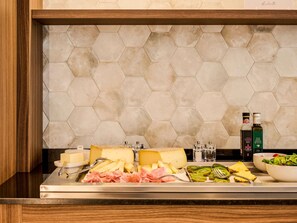 A buffet with cheese, meats, and fruits, and two bottles of olive oil on a counter with a hexagonal tile backsplash.