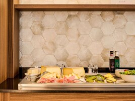 Een buffet met kaas, vleeswaren en fruit, en twee flessen olijfolie op een aanrecht met een betegelde achterwand in zeshoekvorm.