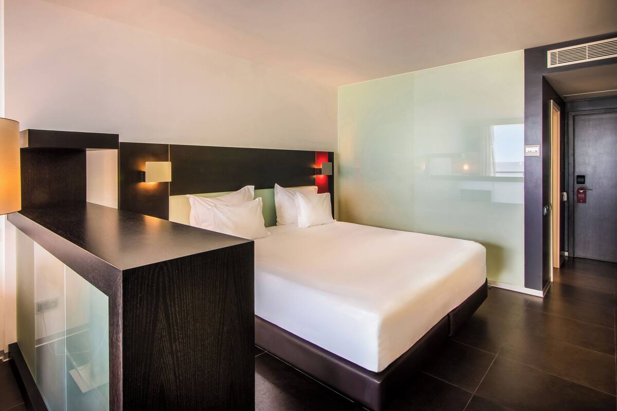 A modern hotel room with a bed, bedside table, lamp, and a wall-mounted light.