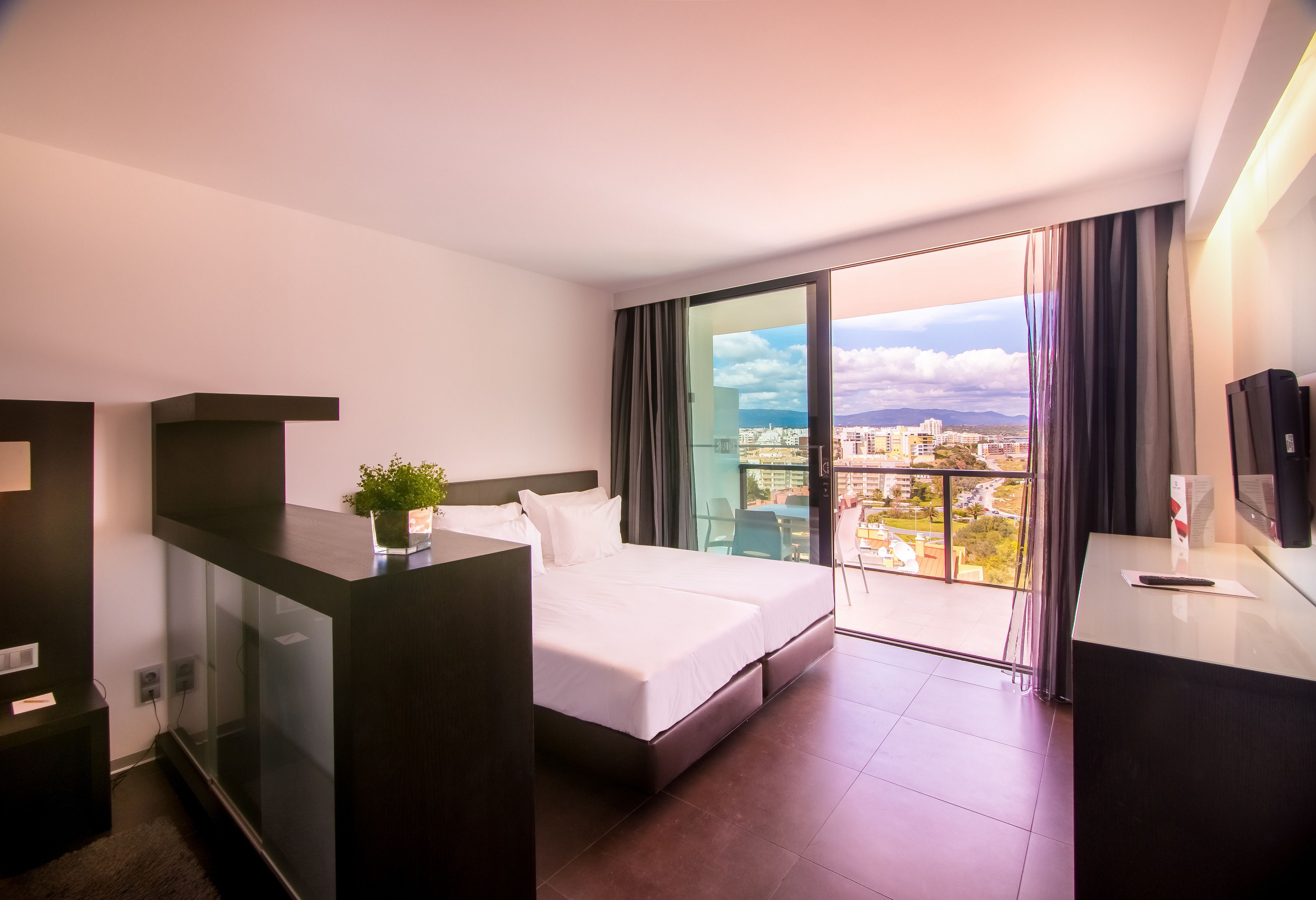 A modern hotel room with a bed, a desk, a glass partition, and a view of the city.