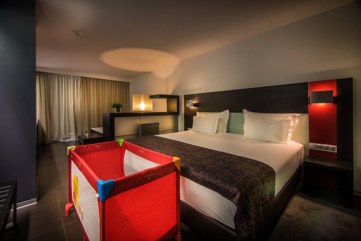 A hotel room with a large bed, a desk with a lamp, a red playpen, and a view of the outside through the window.