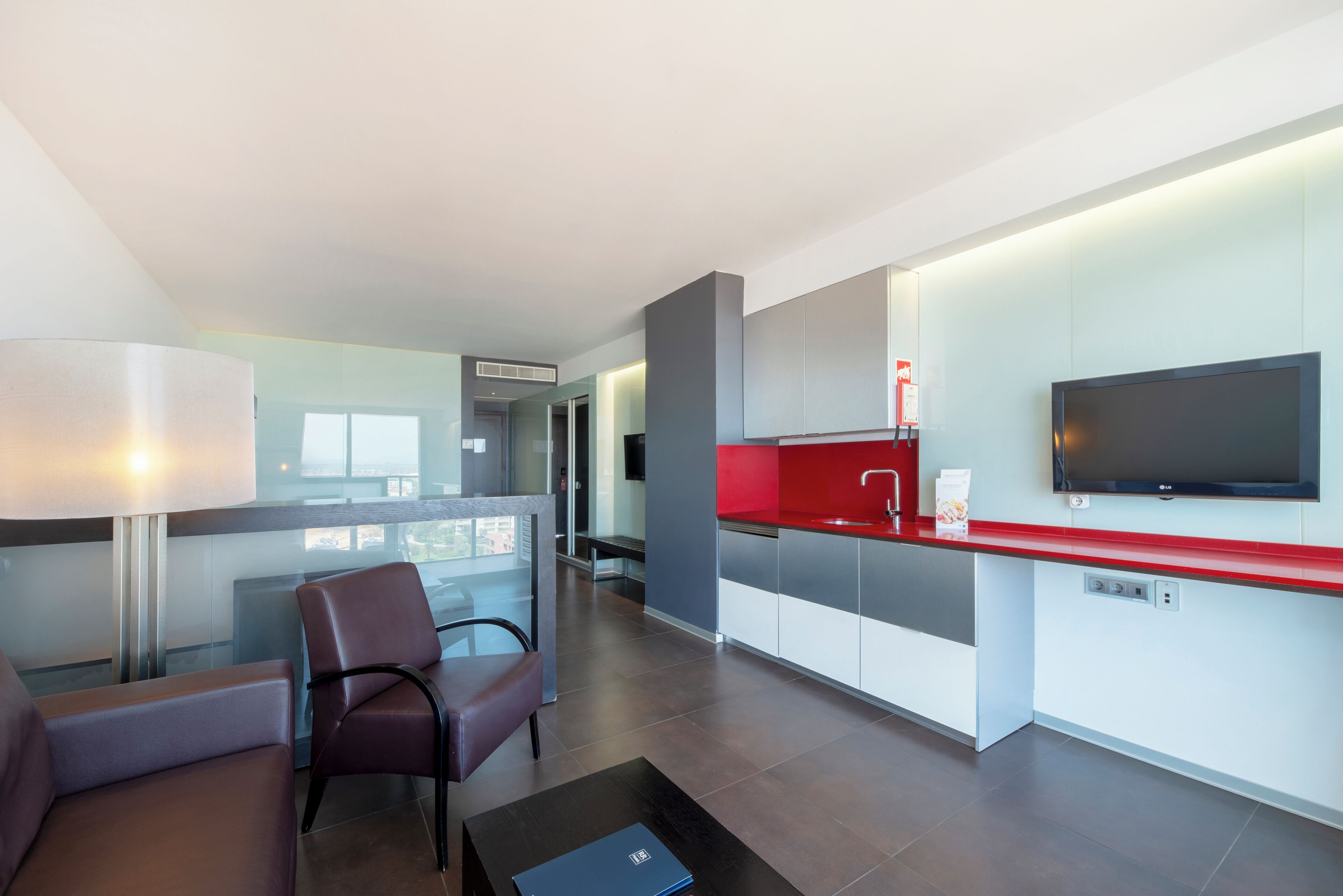 A modern hotel room with a kitchenette, a flat-screen TV, a sofa, and a dining table.