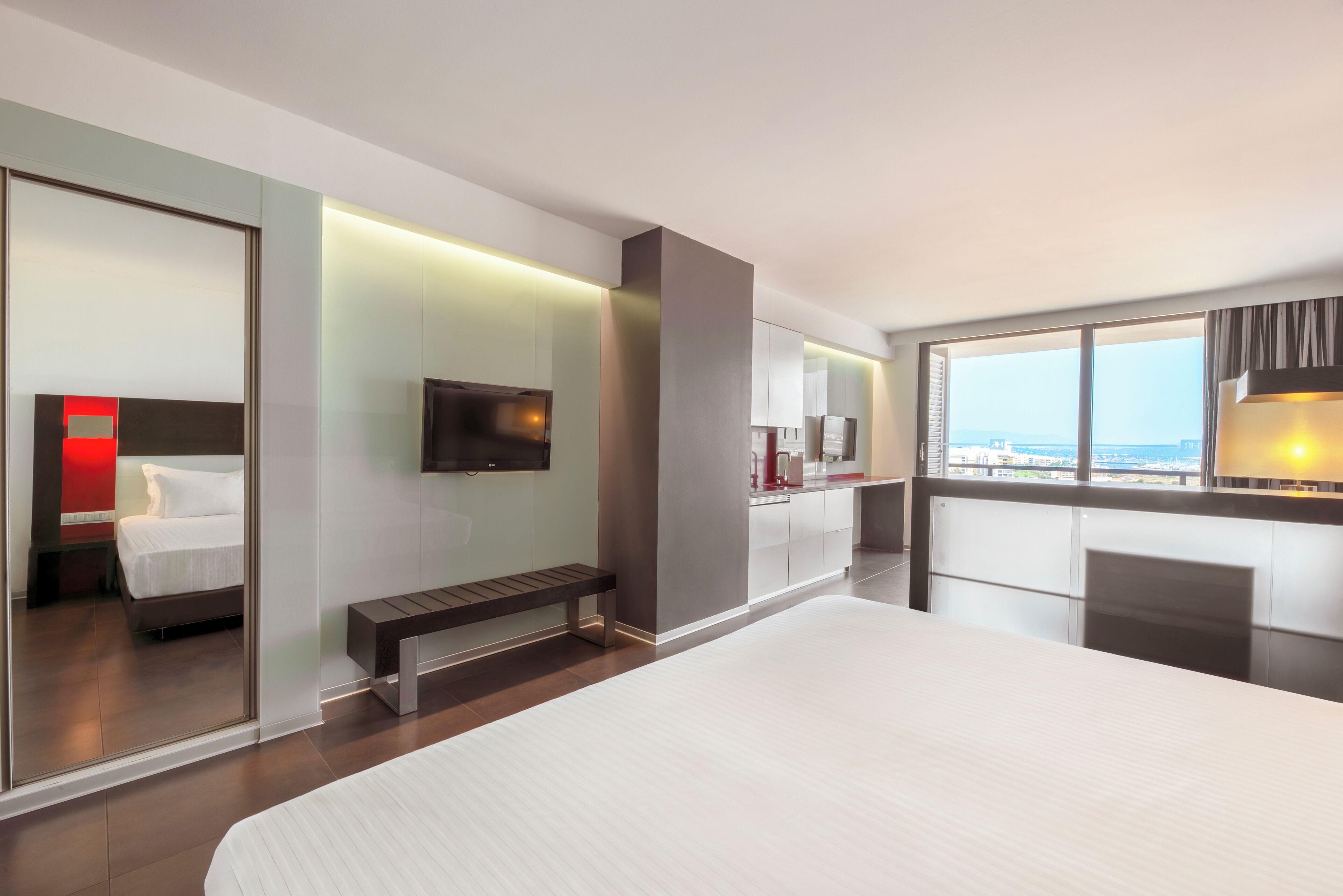 Quarto de hotel moderno com uma cama grande, um banco, uma TV, uma kitchenette e vista para a cidade.