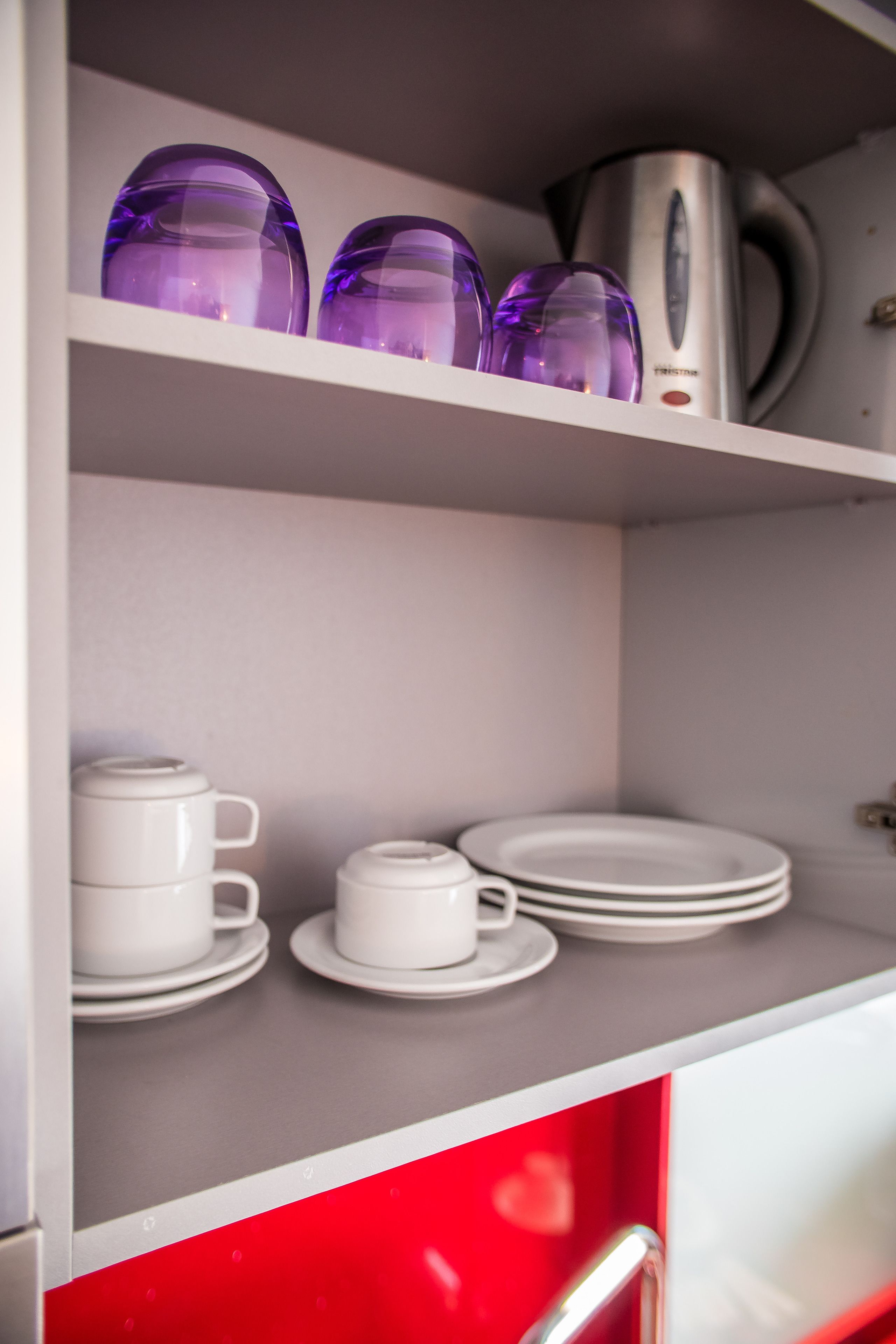 A kitchen with white cabinets, a red lower cabinet, and shelves with purple glassware and white dishes.