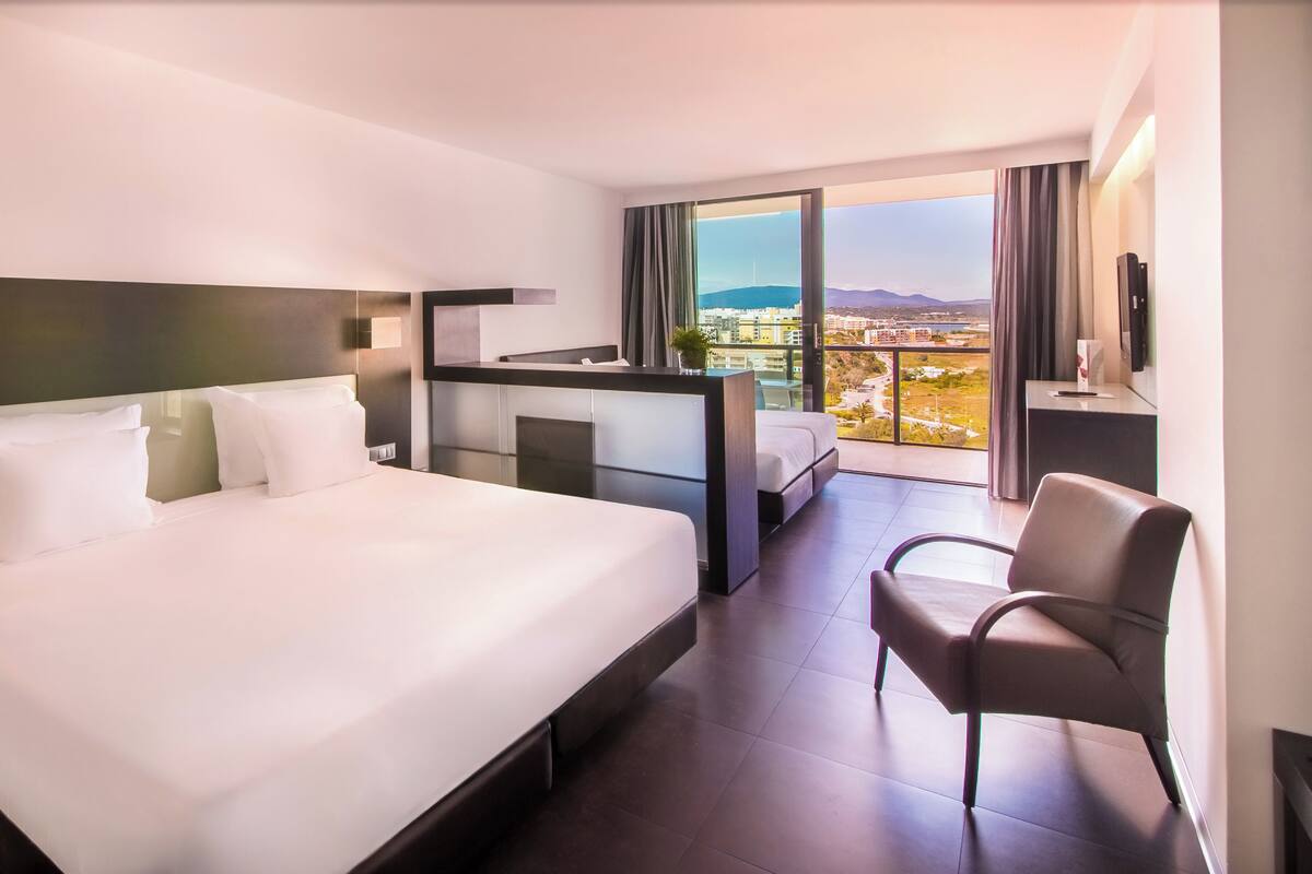 A hotel room with a large bed, a desk, a chair, and a view of the city.