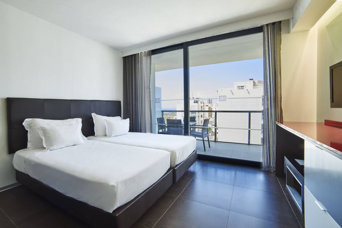 A hotel room with a large bed, a desk, and a balcony with a view of buildings.