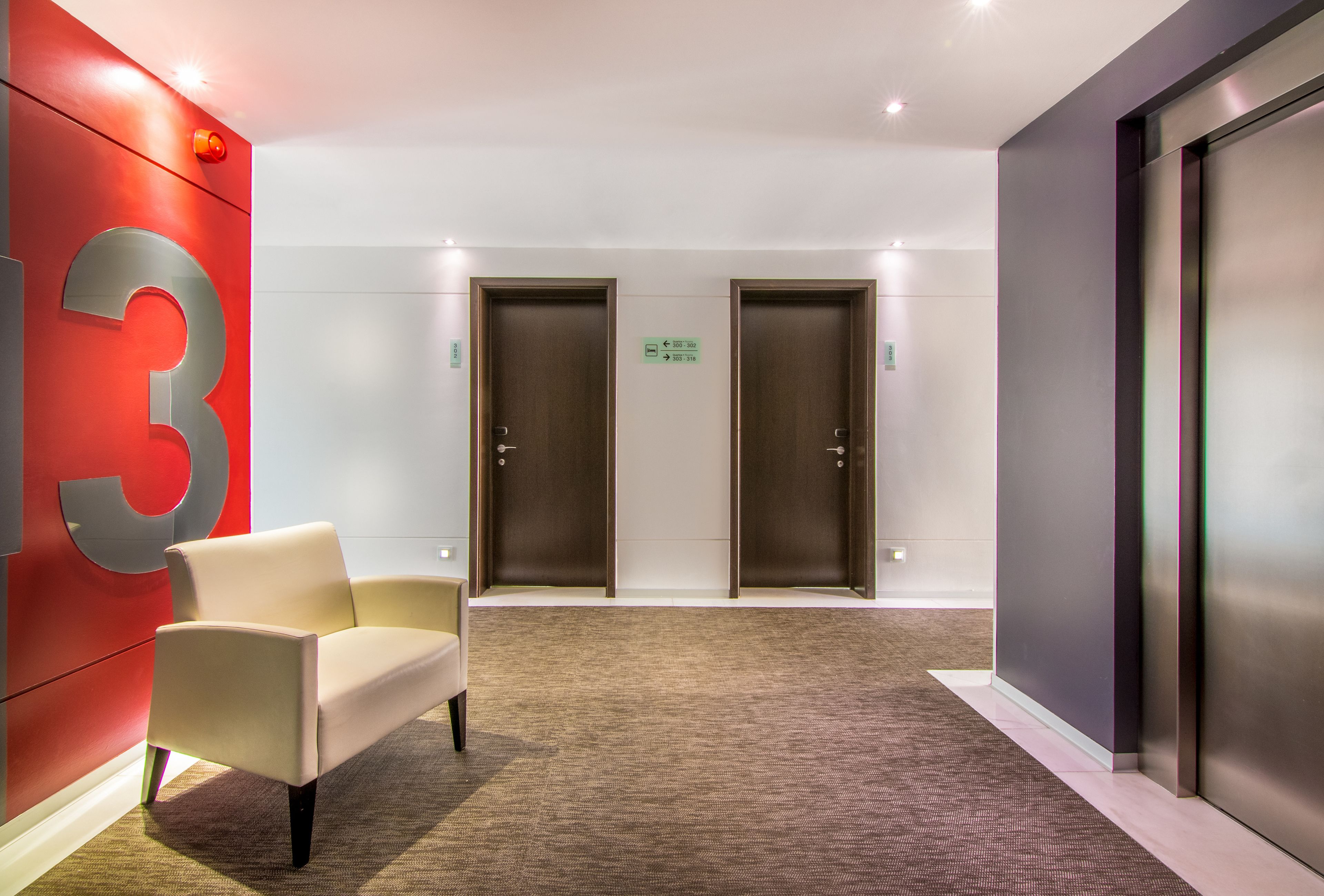 Un lobby moderno con una pared de acento roja, un sofá beige, dos puertas y un ascensor metálico.