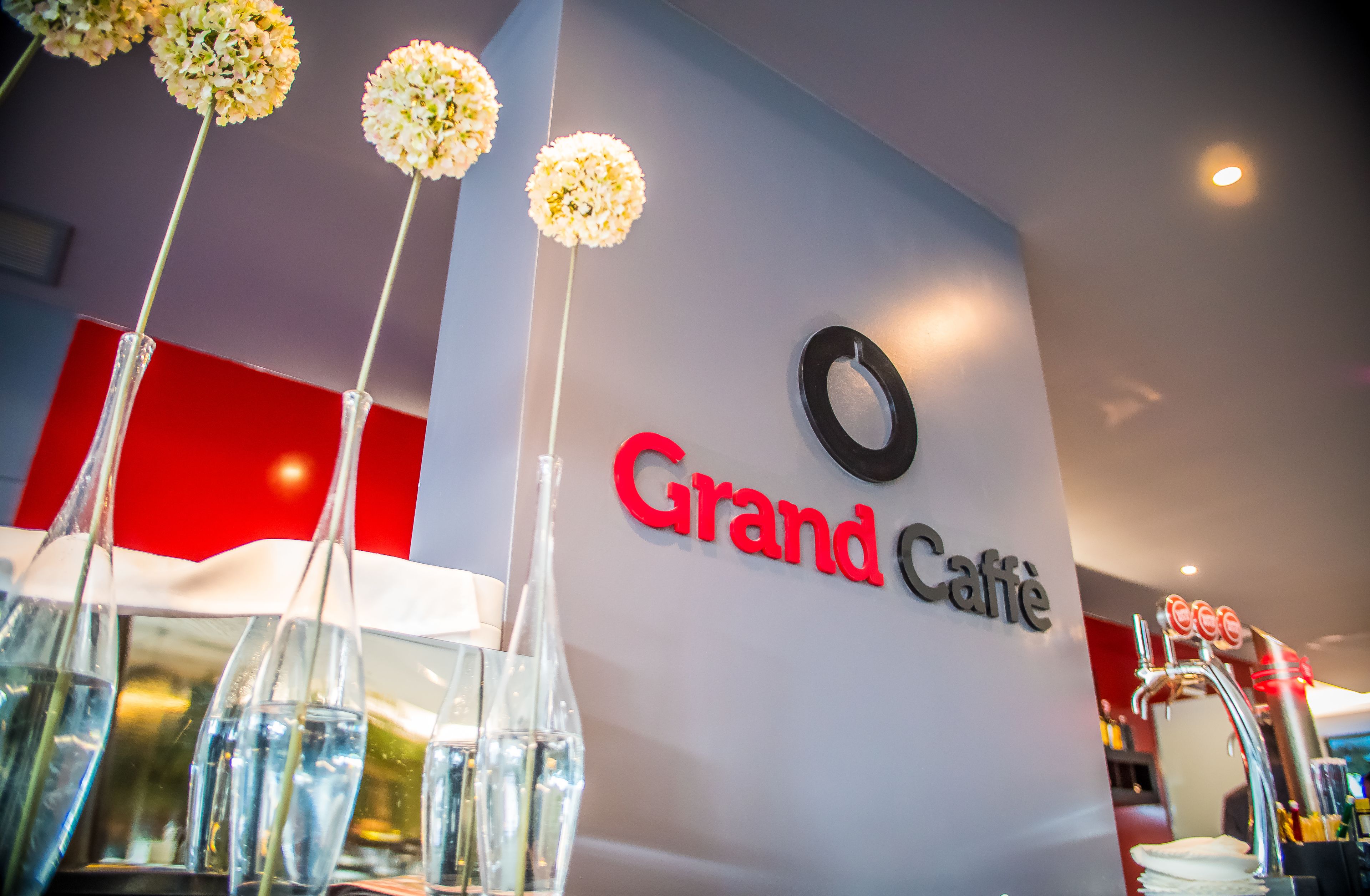 A Grand Caffè sign with a modern design, a bar area with taps, and clear glass vases with flowers.