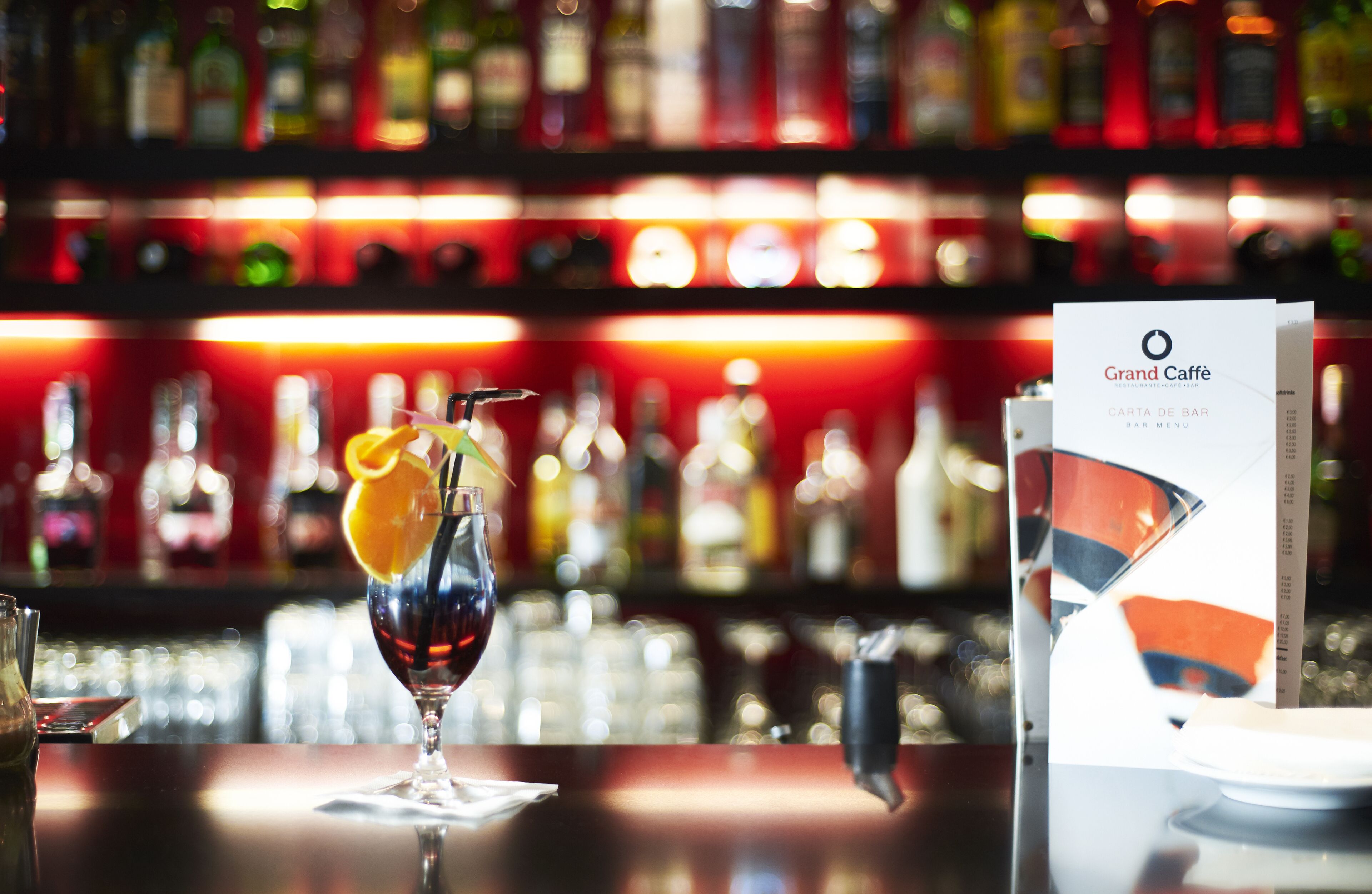 A bar setting with a drink in a glass, a menu, and shelves of bottles in the background.