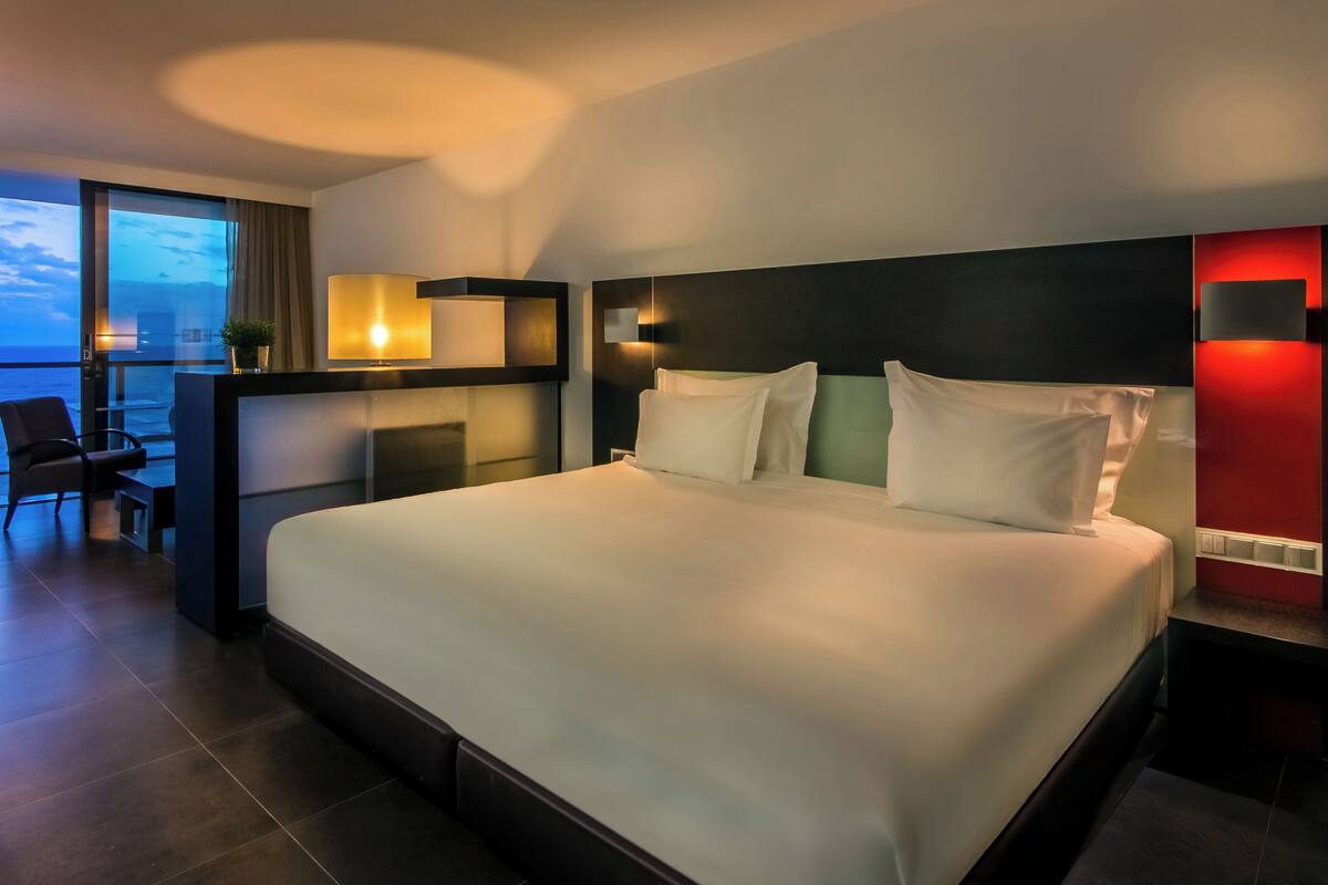 A modern hotel room with a large bed, a desk with a chair, a view of the sea, and a balcony.