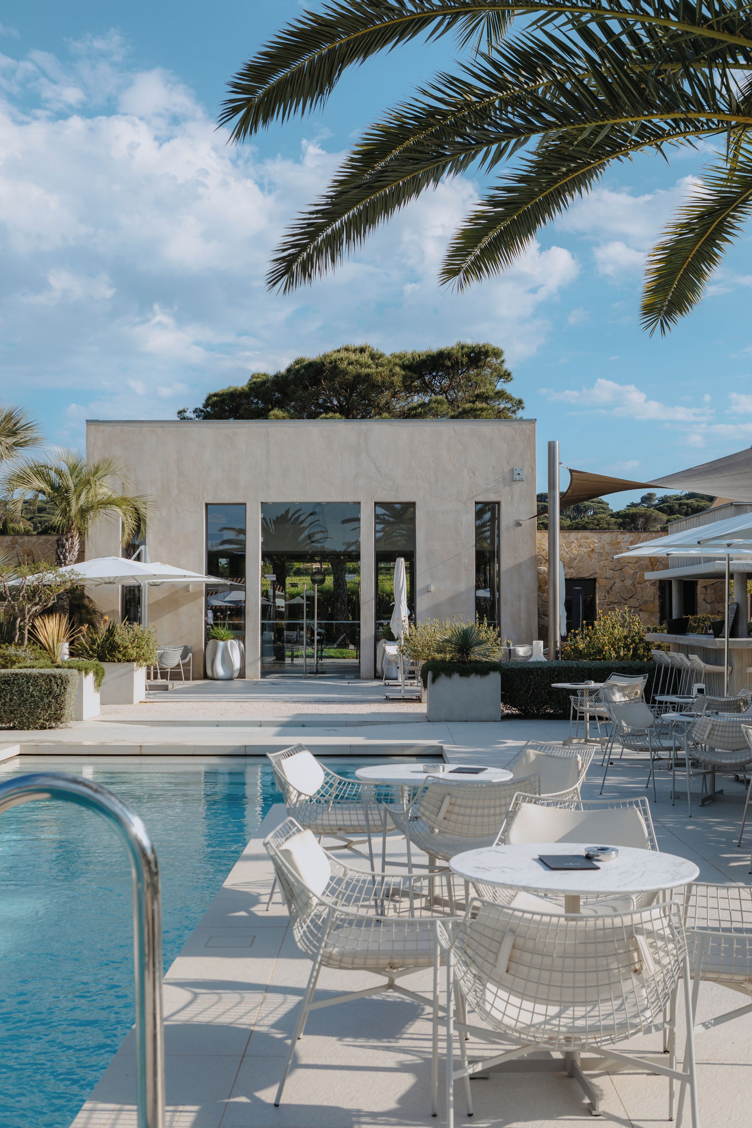 Un espace piscine extérieur moderne, doté de chaises longues et de parasols, un bâtiment en pierre avec de grandes fenêtres et un ciel bleu dégagé.