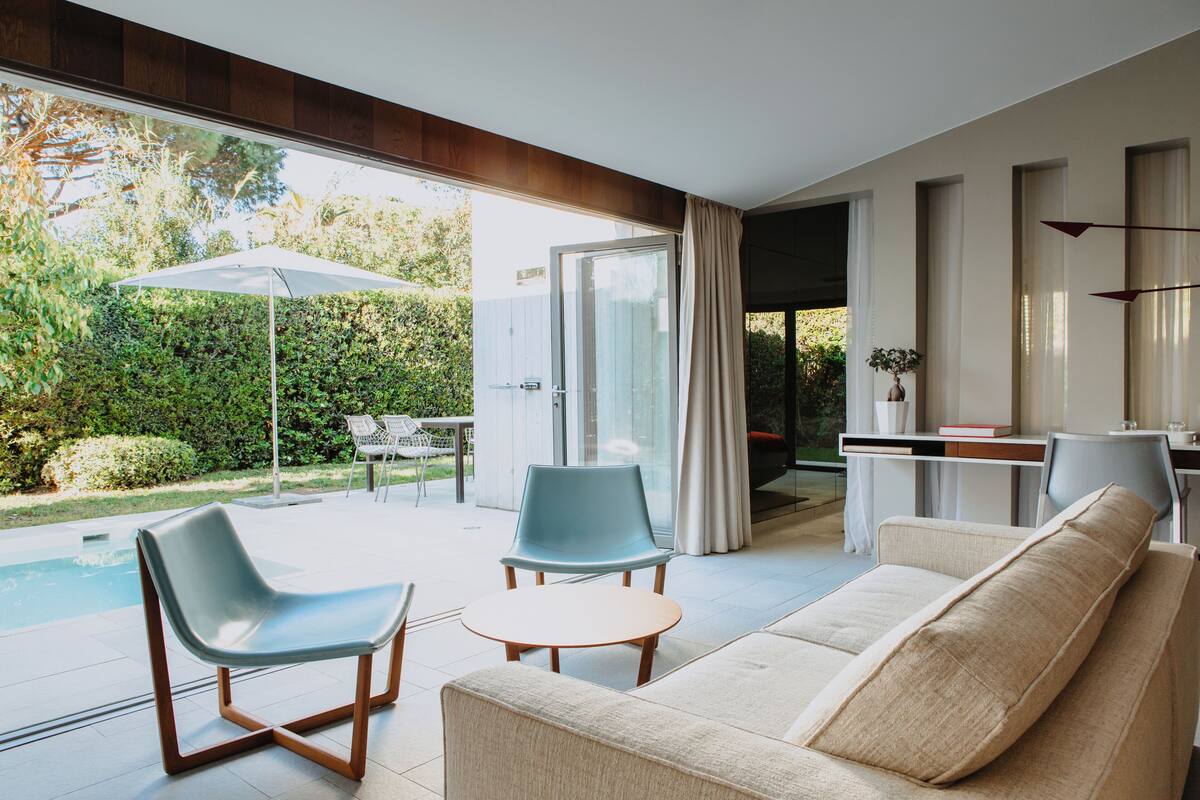 Ein modernes Wohnzimmer mit einer Couch, zwei Sesseln, einem kleinen Tisch und einer Glastür, die auf eine Terrasse mit Pool und Bepflanzung führt.