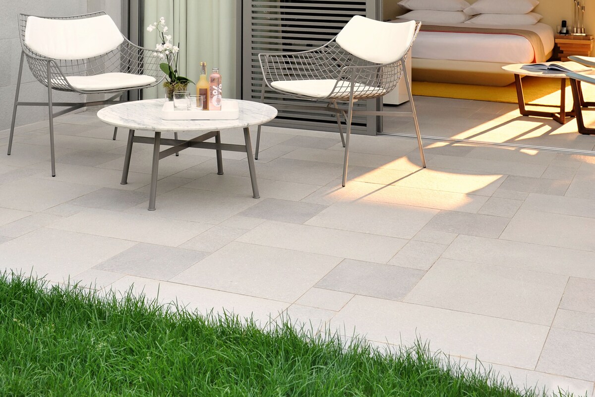 Un patio ext érieur moderne avec deux chaises blanches à structure métallique, une table ronde et un lit doté d’une tête de lit blanche.