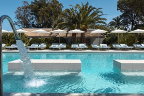 Une piscine entourée de palmiers et d’un bâtiment à toit de tuiles, avec des chaises longues et des parasols à proximité.