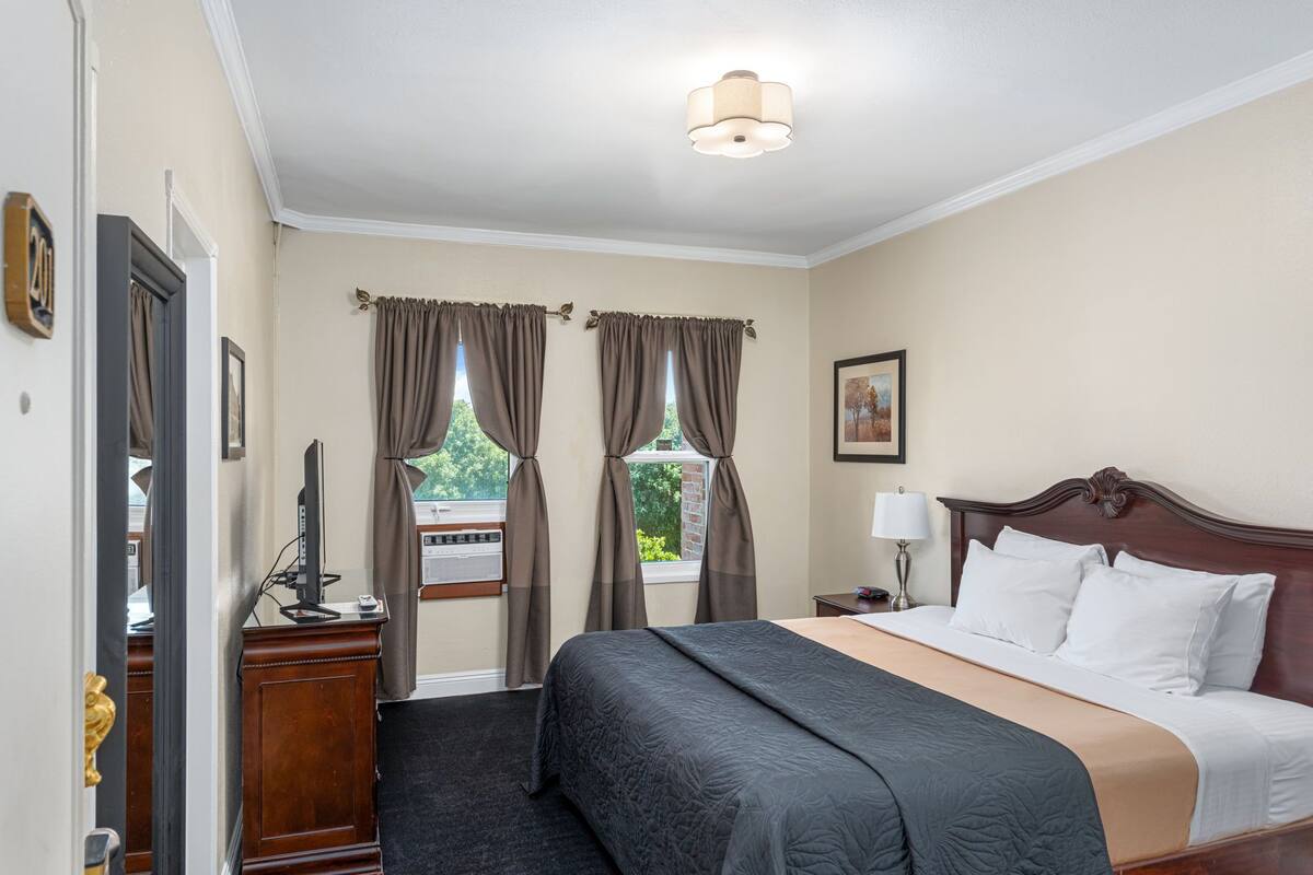 A hotel room with a large bed, a wooden nightstand, a desk with a computer, and a window with curtains.