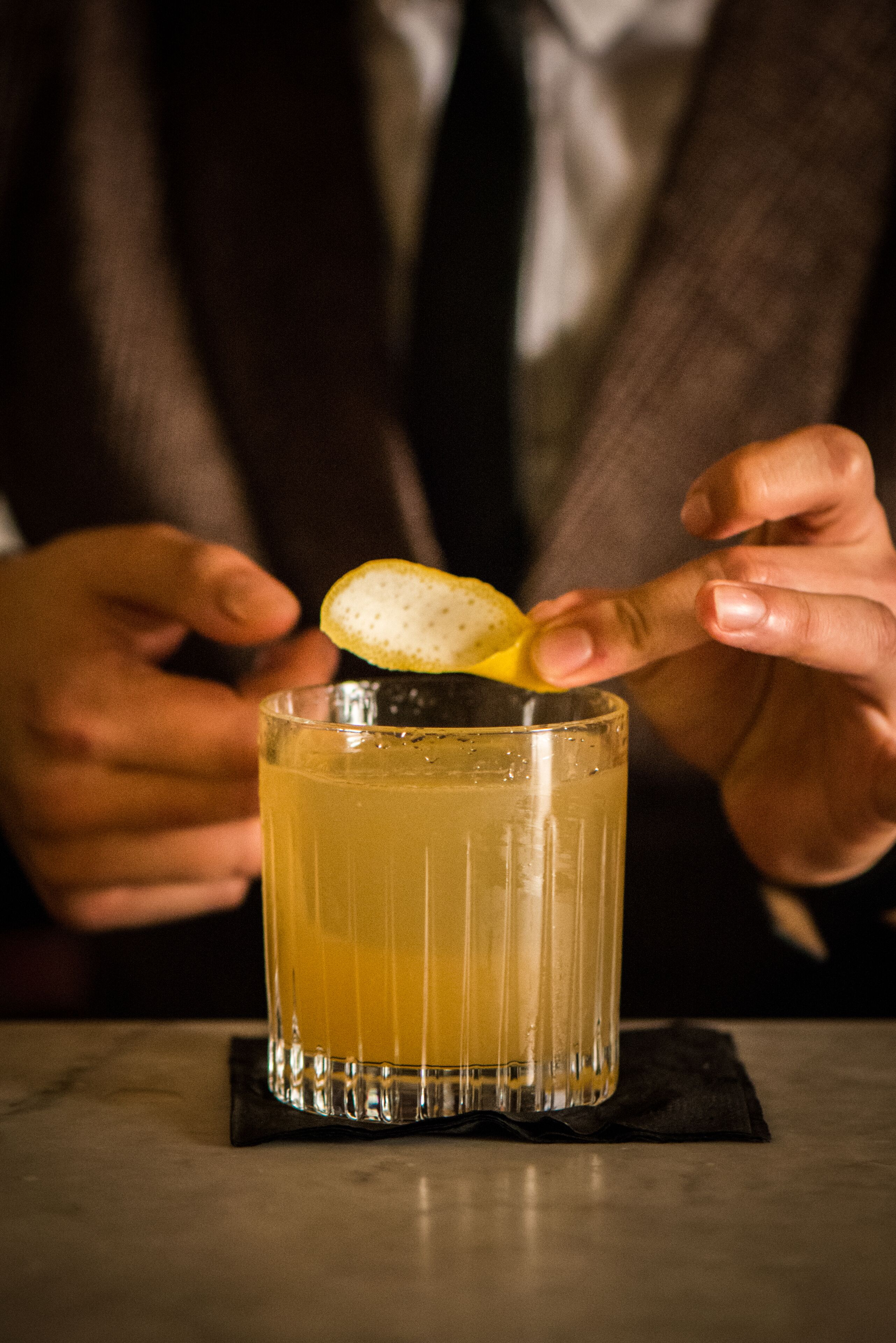 A person squeezing a lemon over a glass of amber liquid, likely a cocktail, on a marble surface.