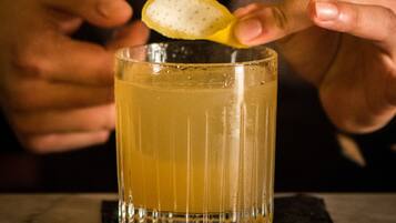 A person squeezing a lemon over a glass of amber liquid, likely a cocktail, on a marble surface.