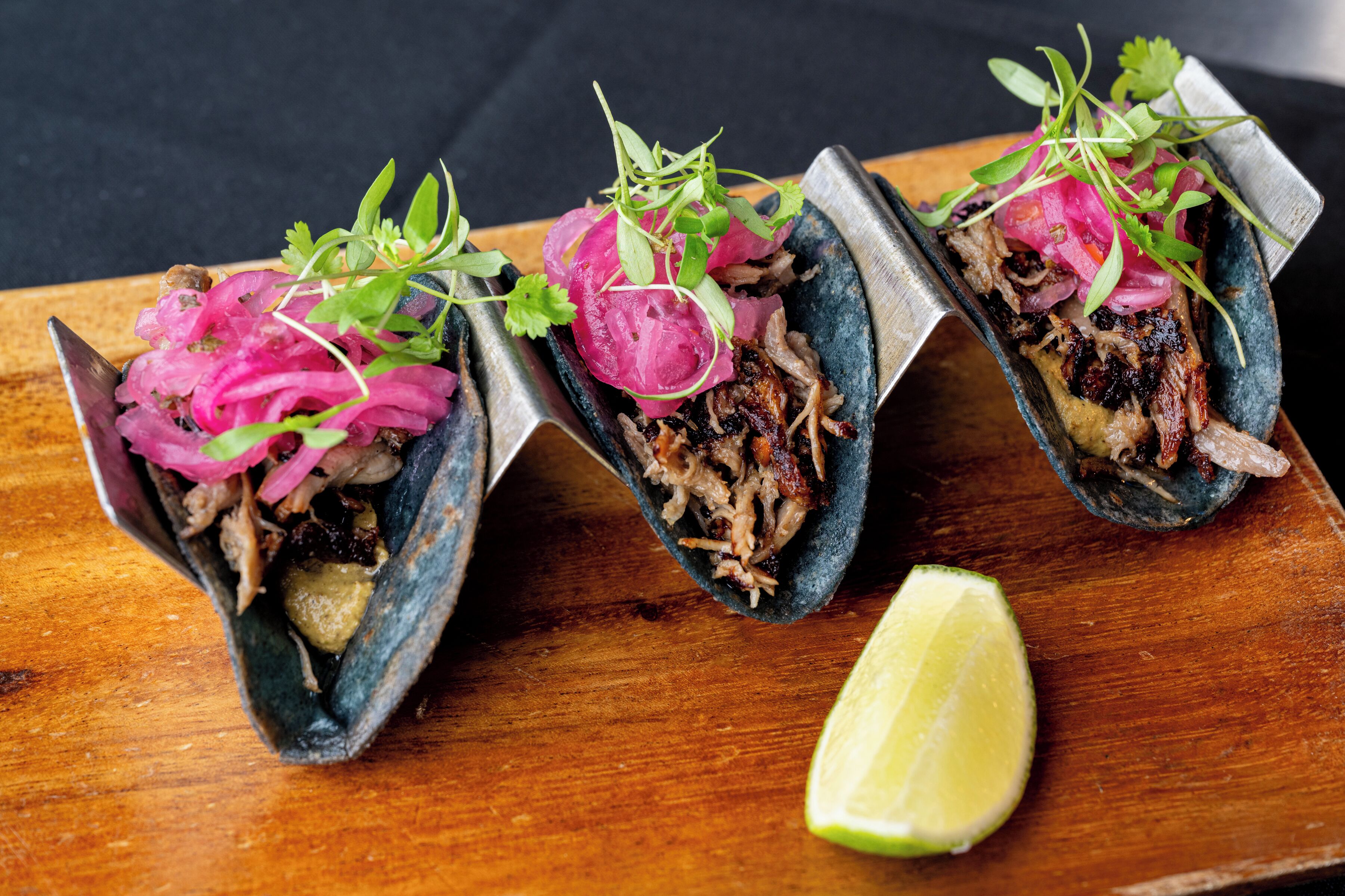 Drie taco's met gescheurd vlees, ingemaakte uien en groen, geserveerd op een houten plank met een partje limoen.