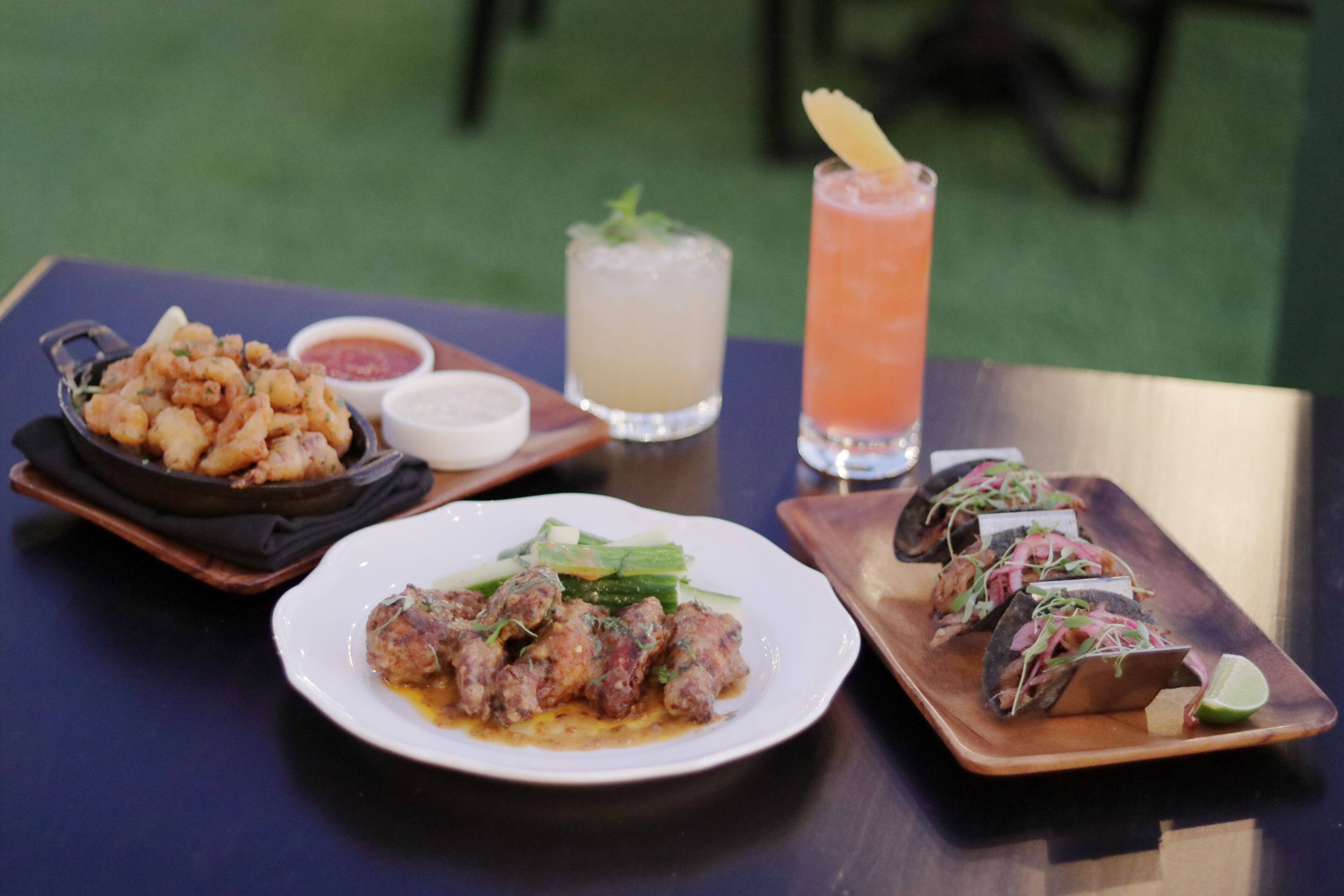 A table with various dishes including fried items, a plate of meatballs, and tacos, along with two drinks.