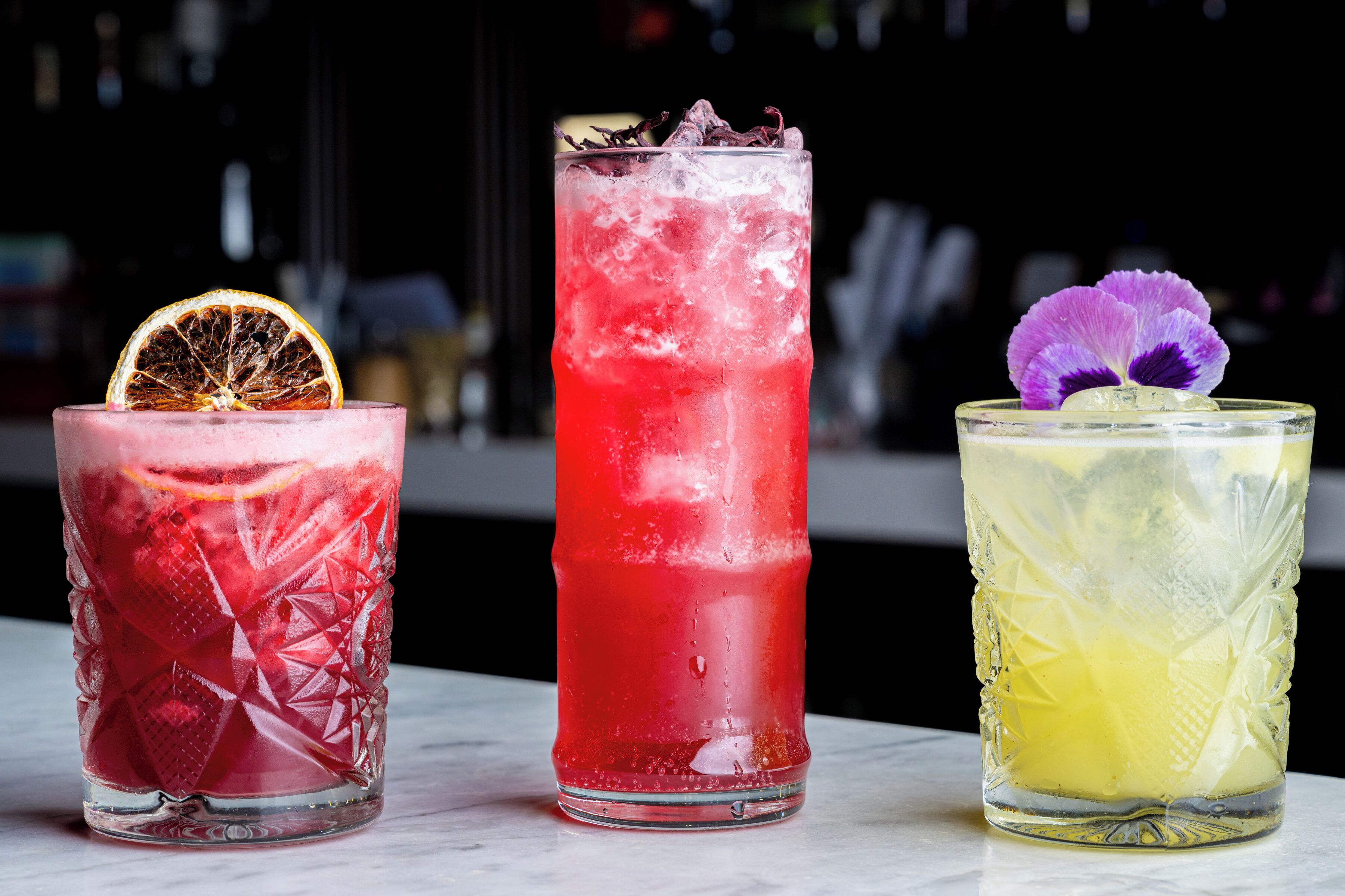 Three glasses of different colored beverages with ice, garnished with a slice of orange and purple flowers.