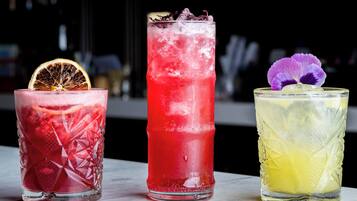 Three glasses of different colored beverages with ice, garnished with a slice of orange and purple flowers.