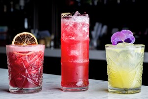 Tres vasos de bebidas de colores distintos, con hielo, adornados con una rodaja de naranja y flores moradas.