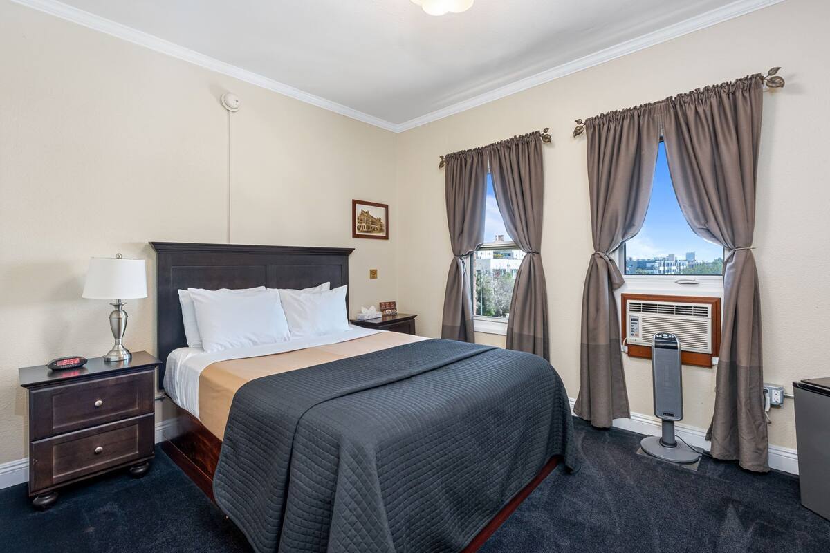 A hotel room with a bed, a nightstand, a lamp, a window with curtains, and an air conditioning unit.