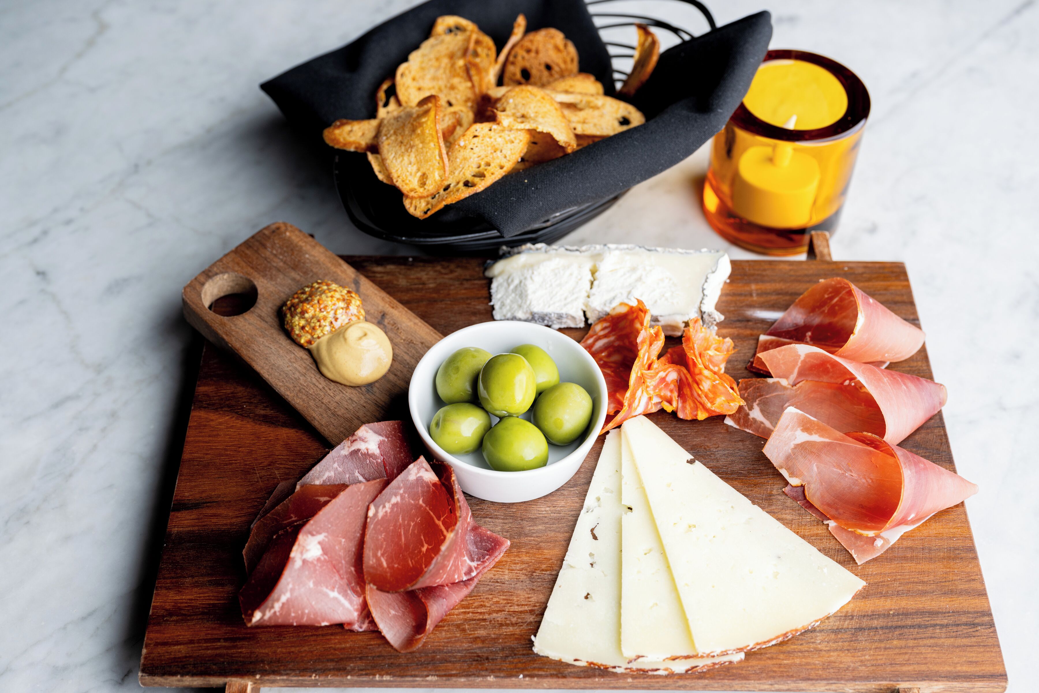 A wooden board with various cheeses, cured meats, olives, and crackers.