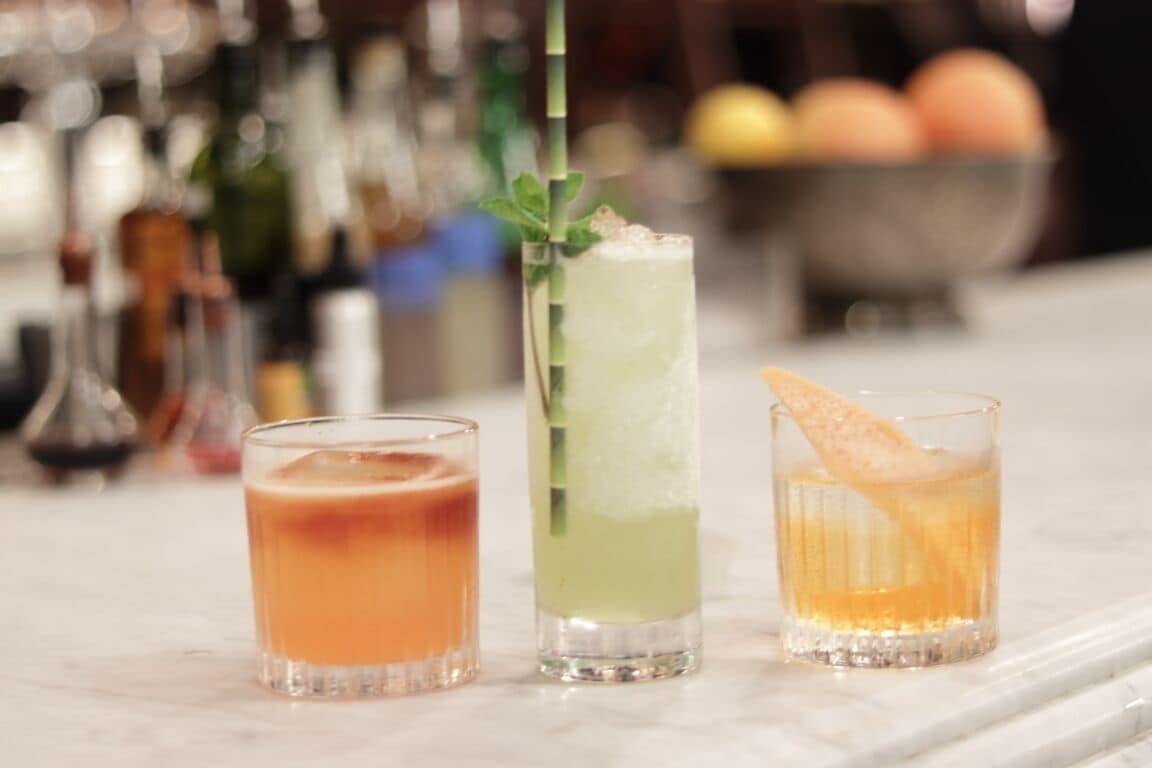 Three cocktails on a bar counter with a blurred background of bottles and a fruit bowl.