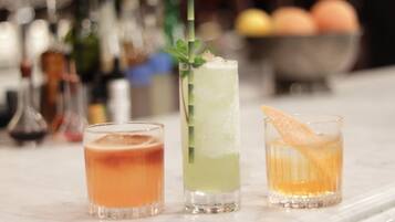 Three cocktails on a bar counter with a blurred background of bottles and a fruit bowl.