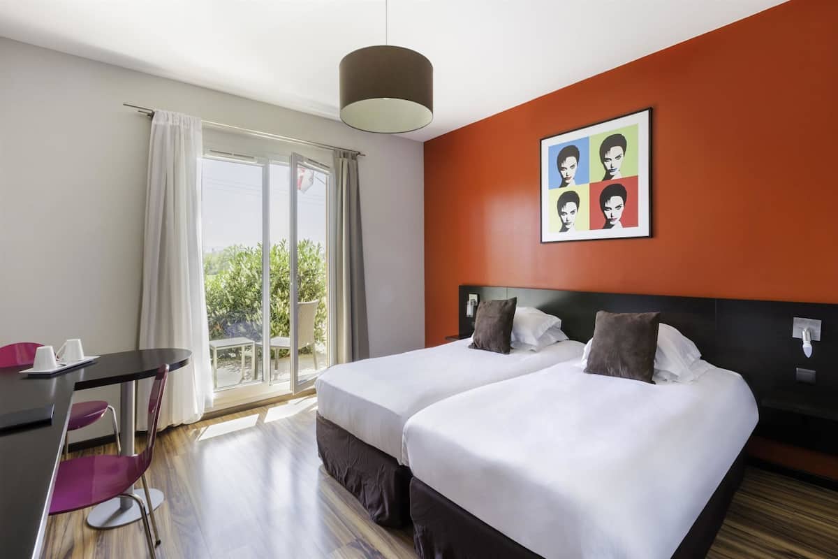 A hotel room with two beds, a desk with chairs, a large window with curtains, and a colorful artwork on the wall.