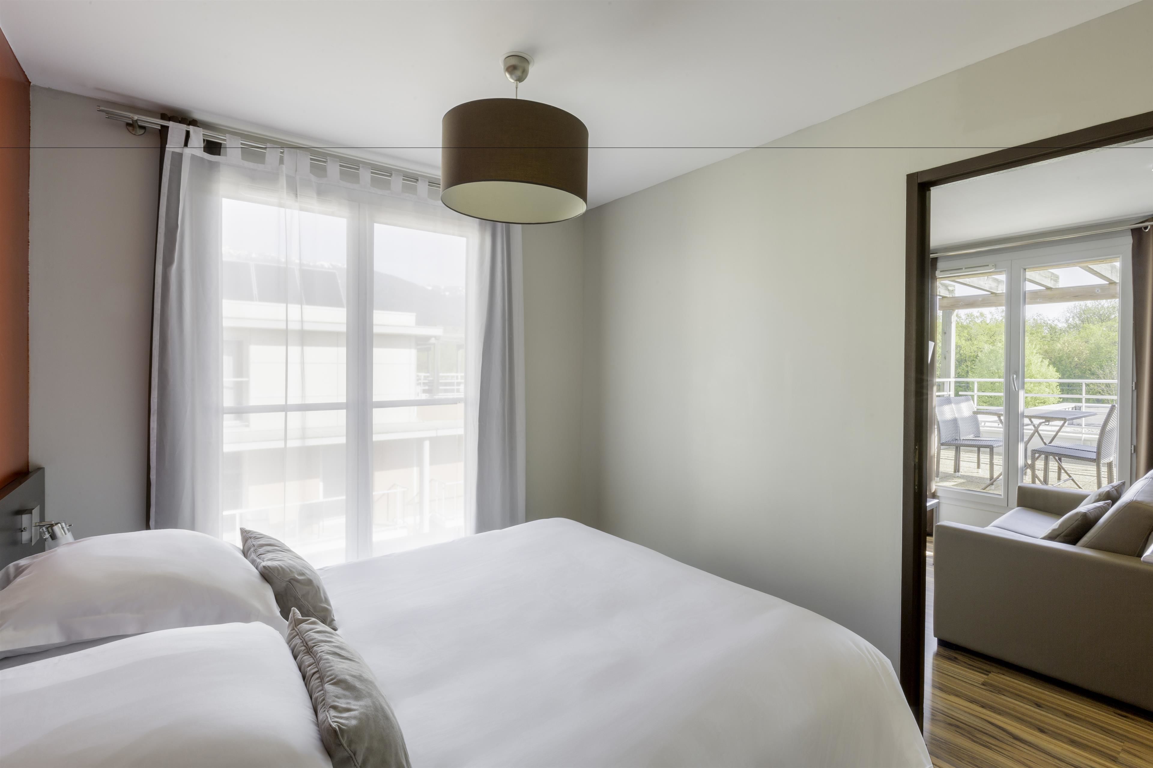A modern hotel room with a large bed, a ceiling lamp, a view of the outdoor seating area through a glass door.