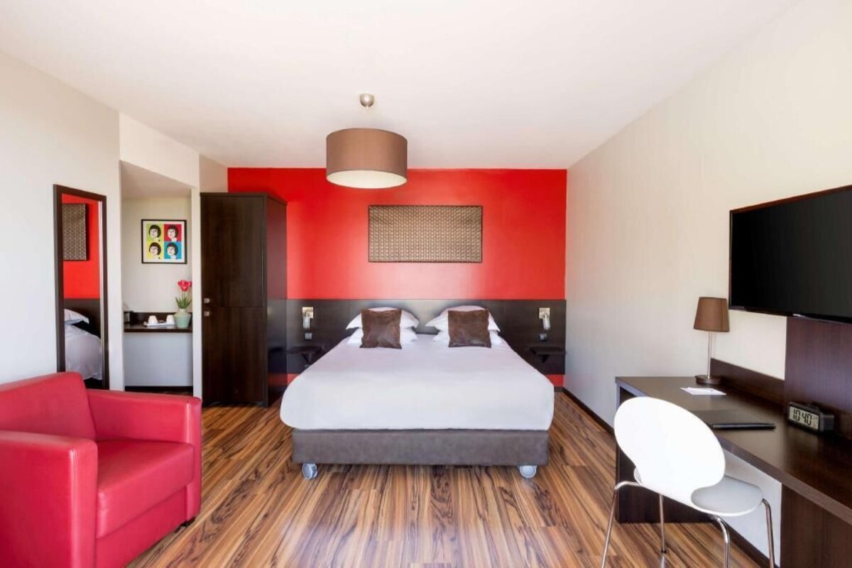 A hotel room with a bed, a desk, a TV, a red chair, and a red wall.