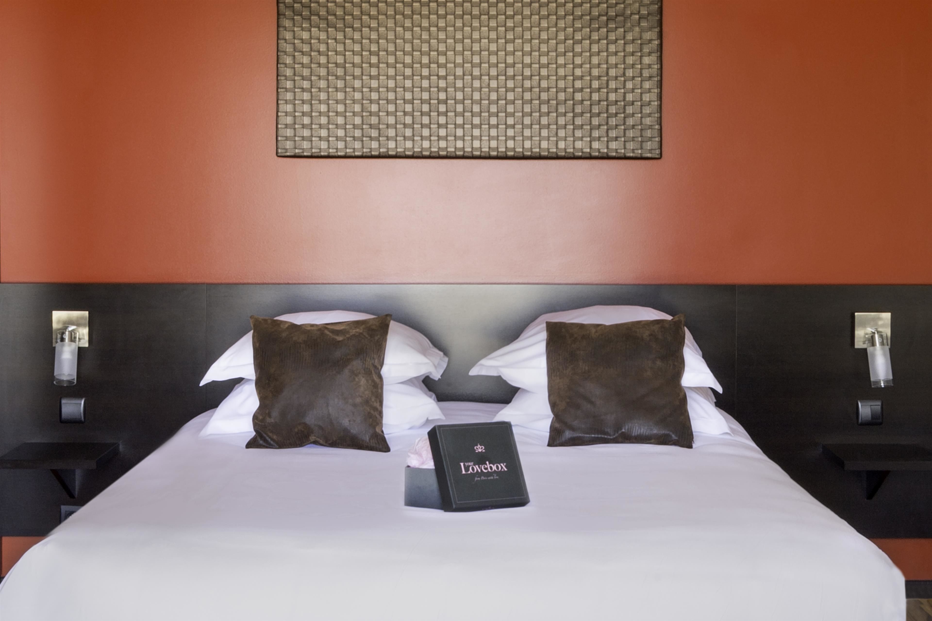 A neatly made bed with a white comforter, two brown pillows, and a small black box labeled "Lovebox" on the bed.