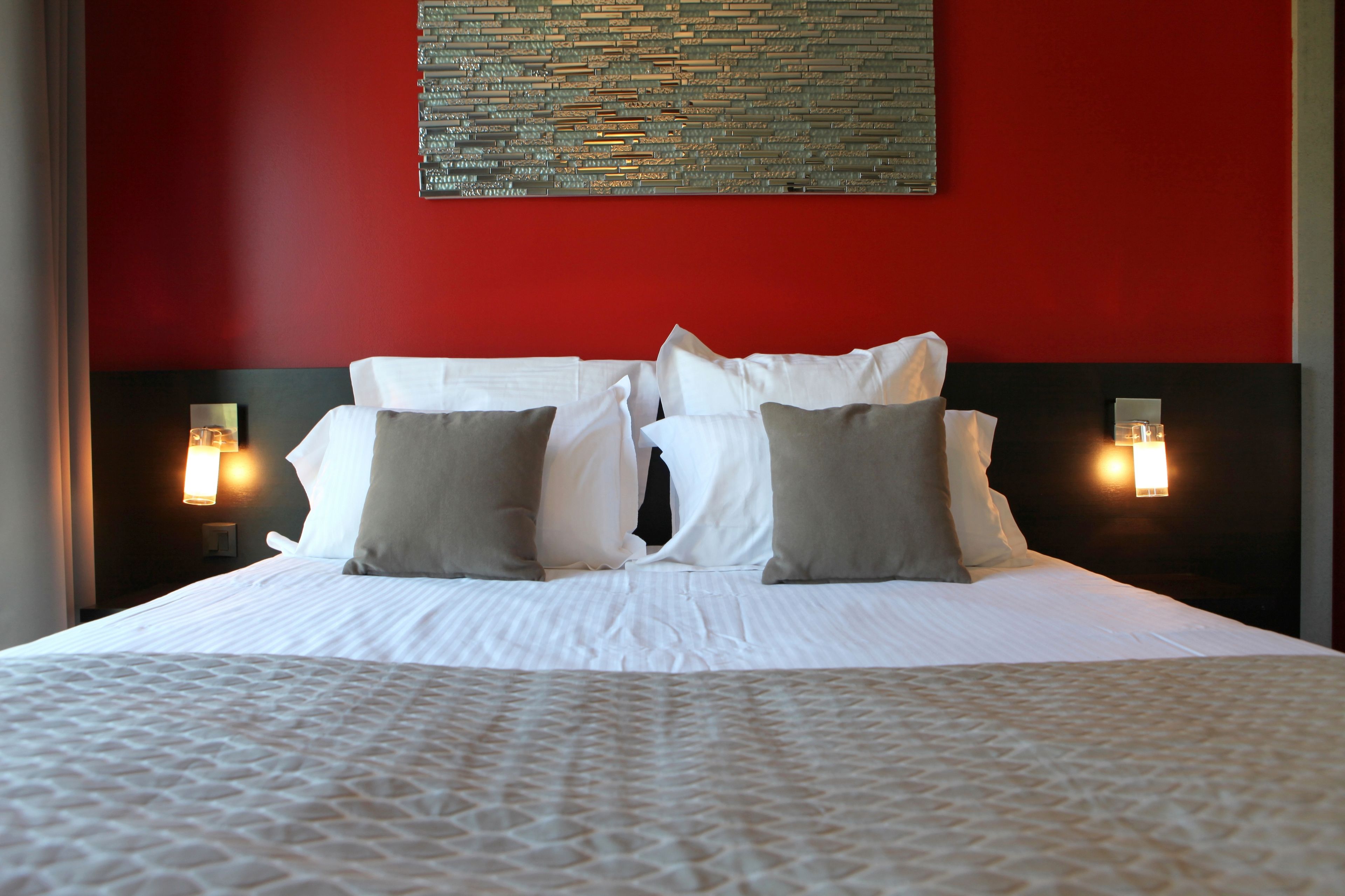A hotel room with a red accent wall, a large bed with white bedding, and two wall-mounted reading lamps.