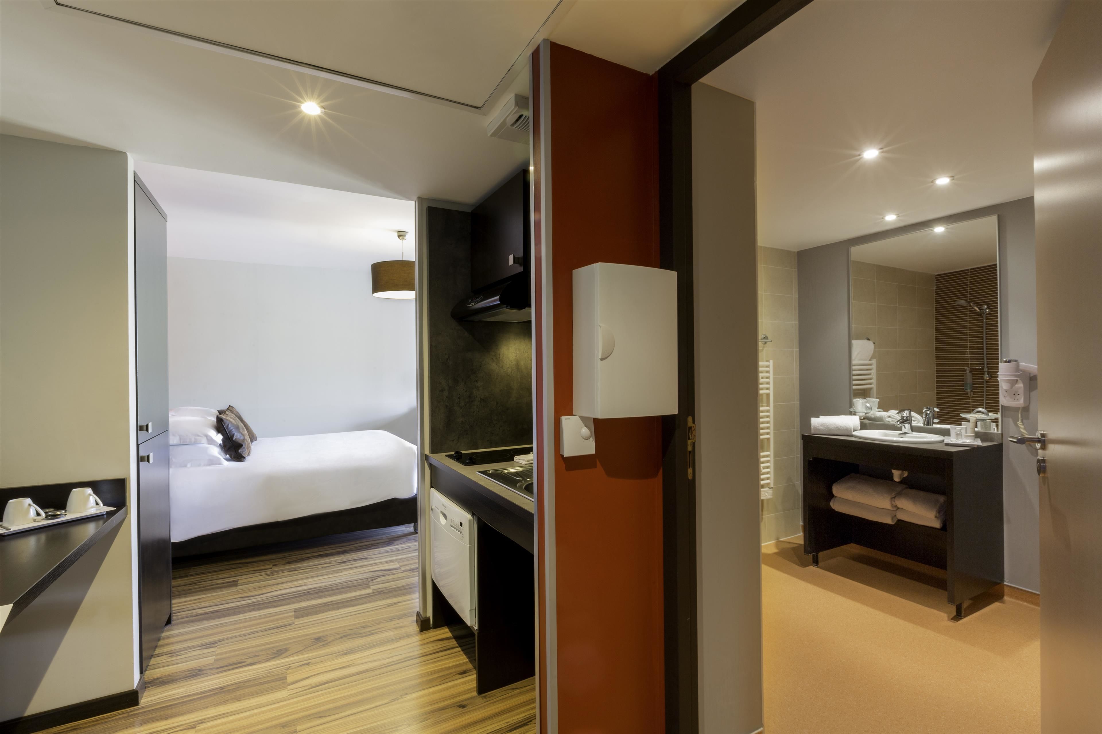 A modern hotel room with a bed, a small kitchenette, a bathroom with a sink and mirror, and a wooden floor.