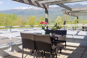 Um terraço com cadeiras de vime, uma mesa, um vaso com flores e vista para montanhas cobertas de neve.