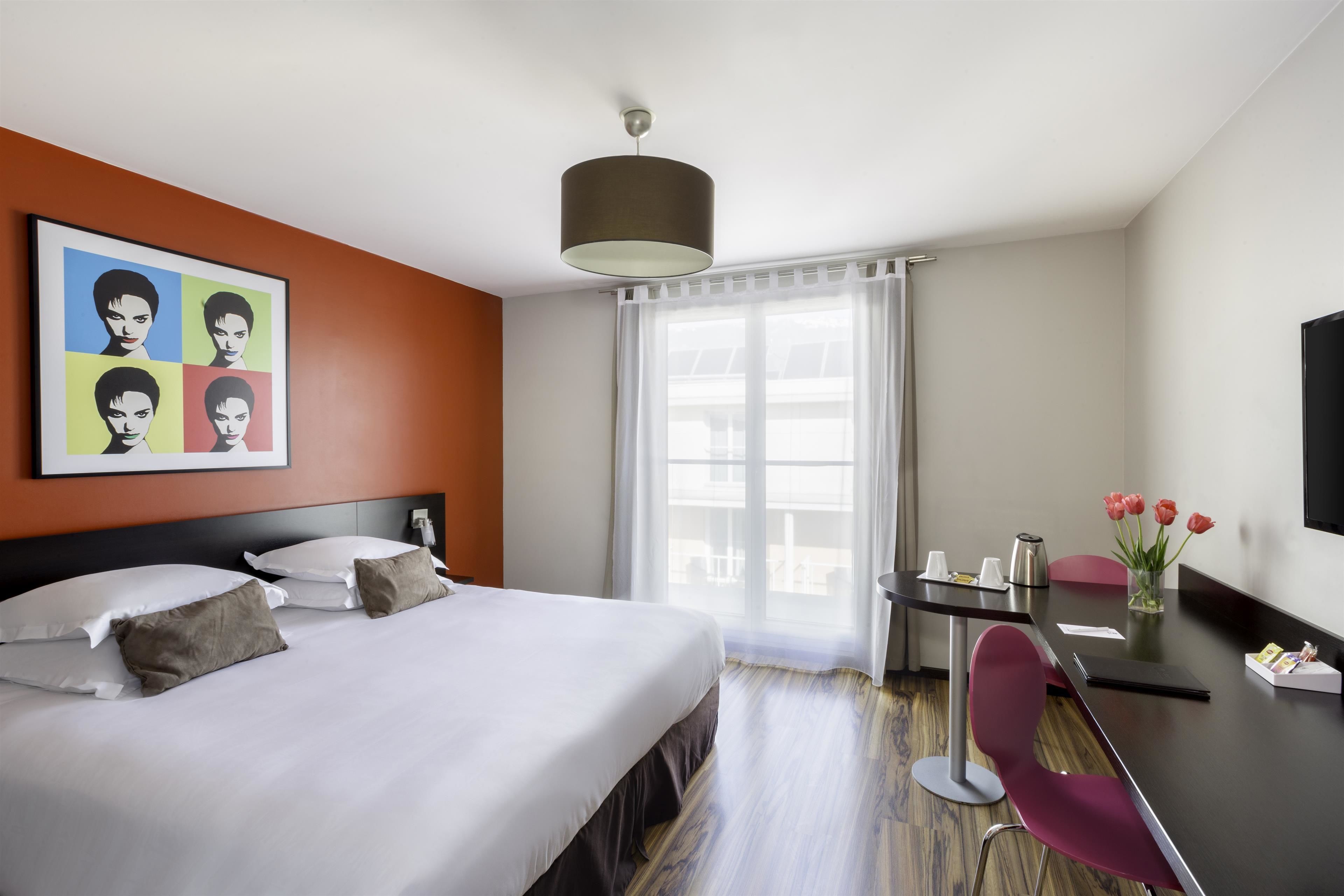 Habitación de hotel con cama, escritorio, silla, un jarrón con flores y un cuadro en la pared.