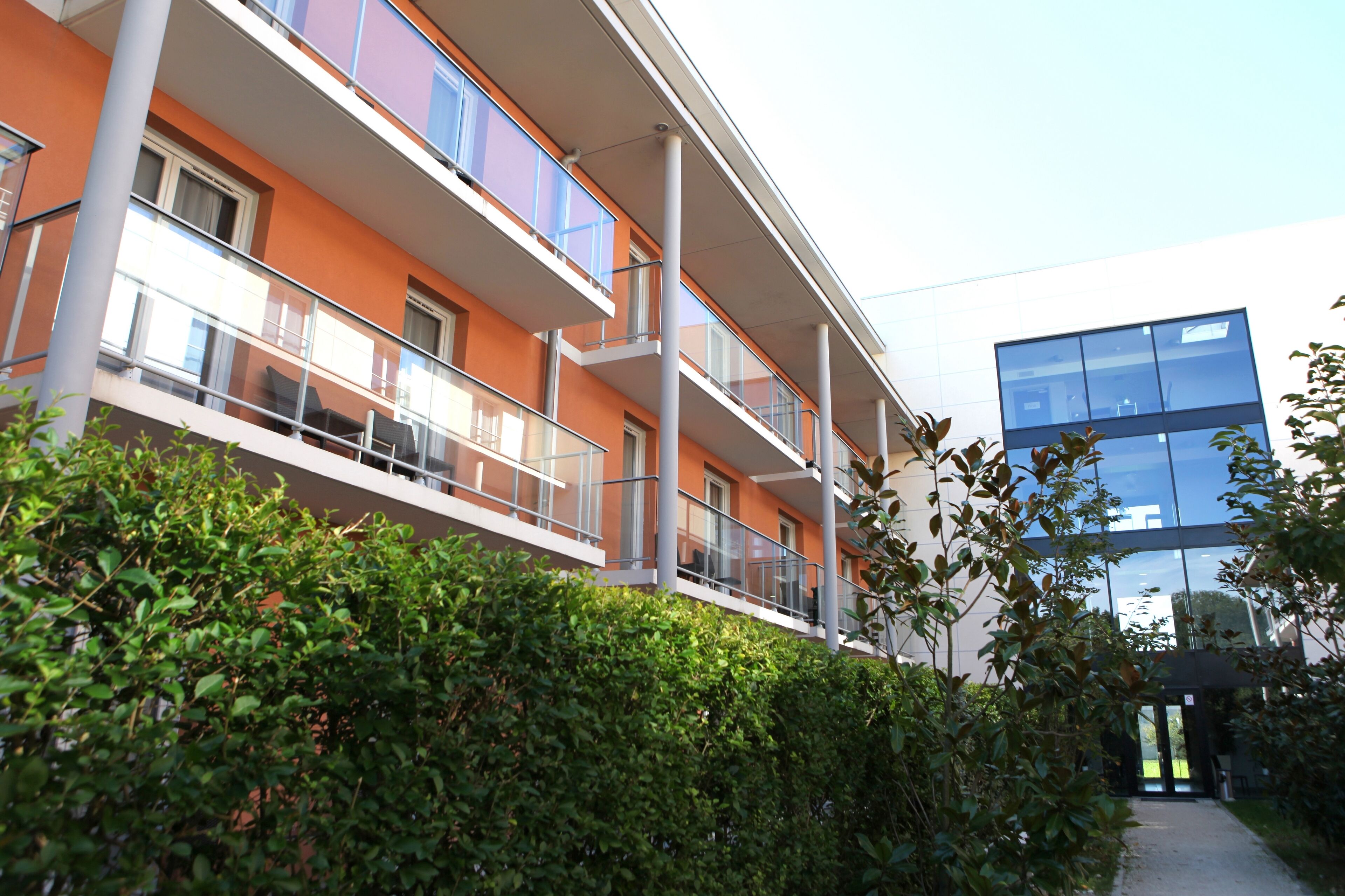 Un edificio de varios pisos con balcones, fachada de vidrio y un camino que conduce a la entrada.
