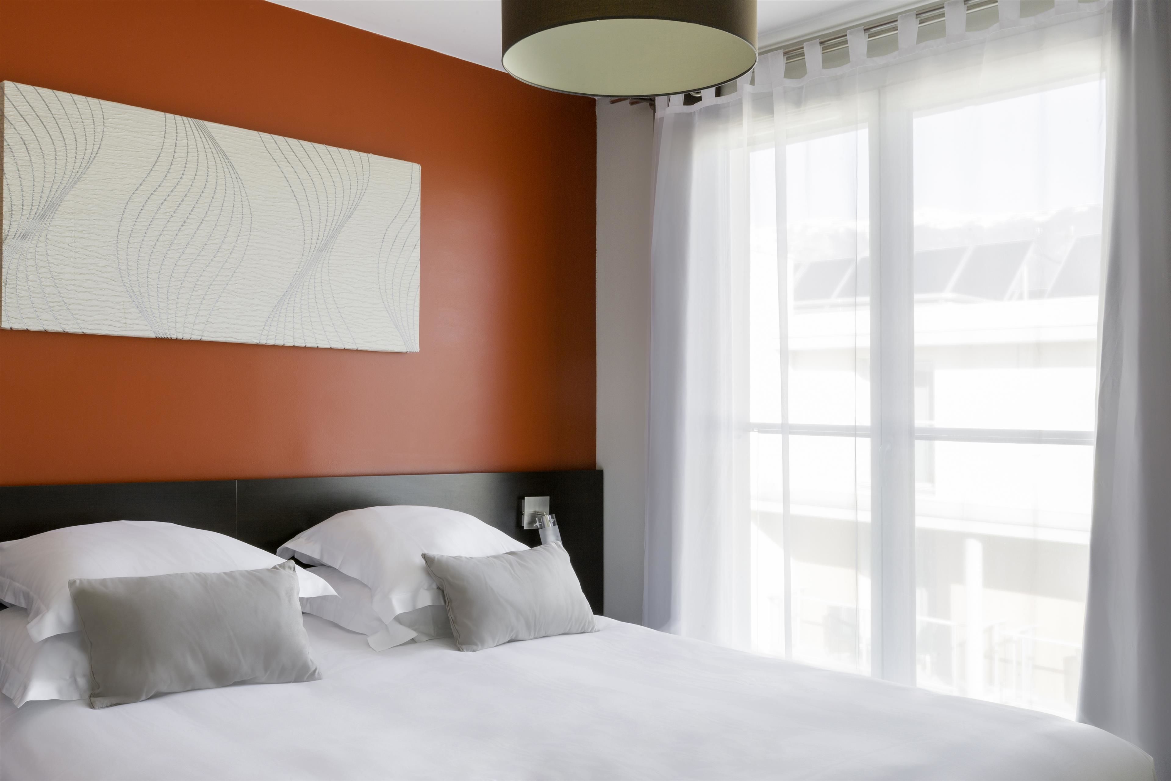 A hotel room with a large bed, white bedding, a modern headboard, and a large window with sheer curtains.
