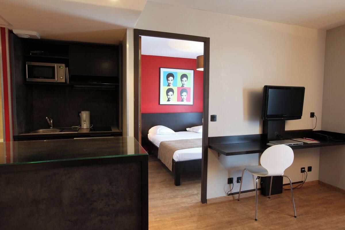 A compact hotel room with a kitchenette, a bed, a desk with a chair, and a television.